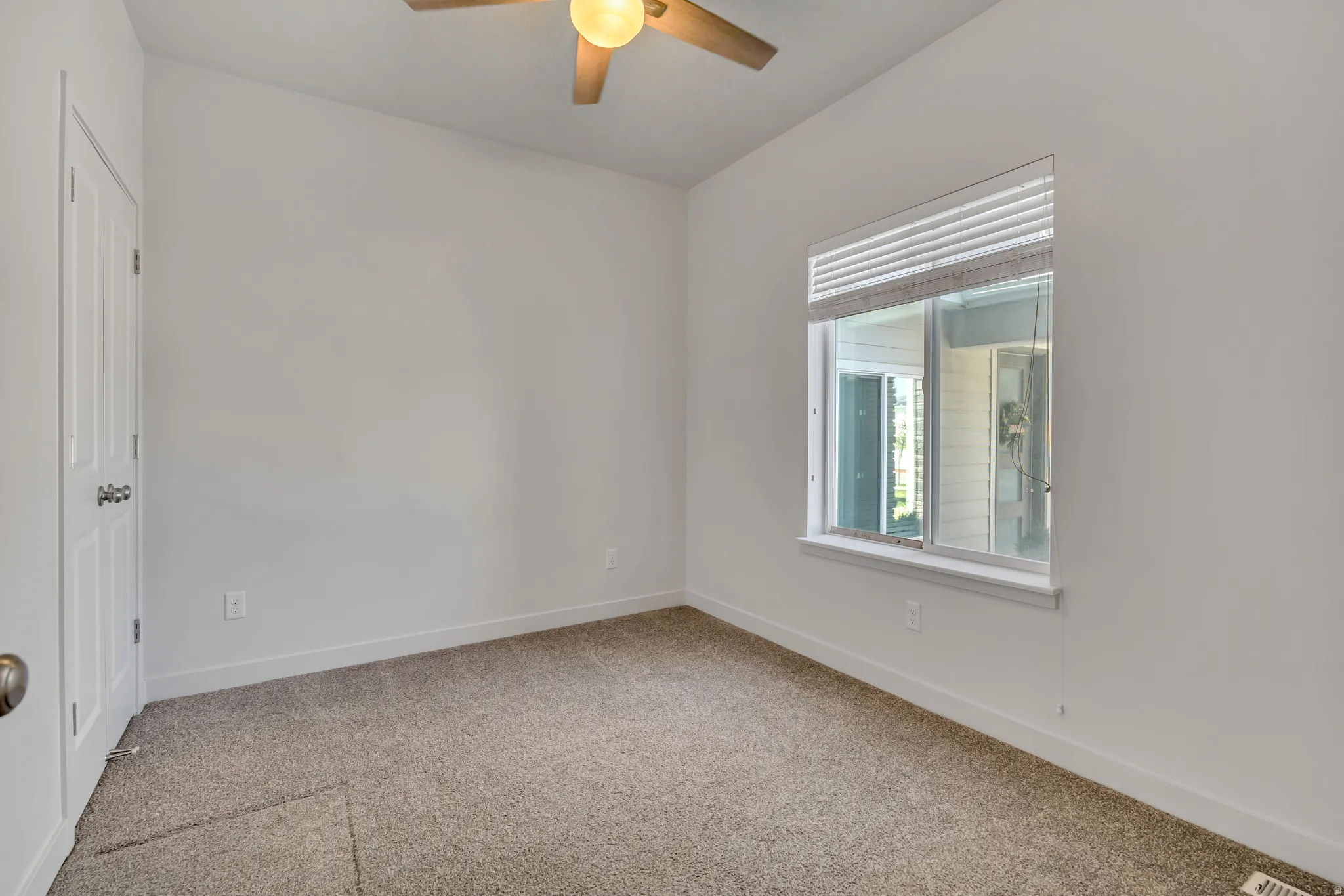 Bright room featuring neutral carpeting, white walls, a large window with blinds, a ceiling fan with integrated lighting, and a white paneled door