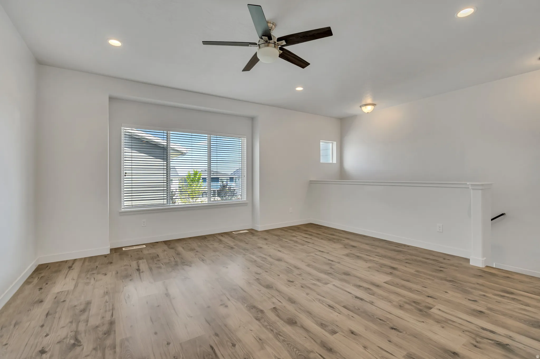 Spacious room featuring wood-finish flooring, a large multi-pane window with blinds, recessed ceiling lighting, and a dark-bladed ceiling fan