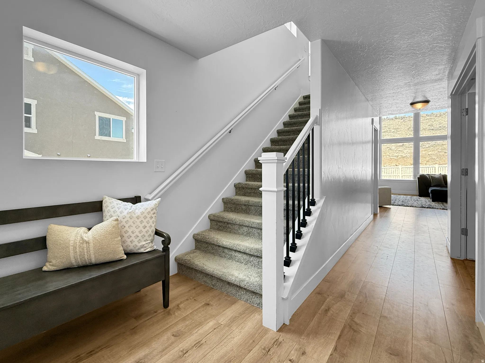 Entryway featuring wood-finish flooring, a carpeted staircase with white risers and black balusters, a large picture window, and a hallway leading to a room with expansive windows