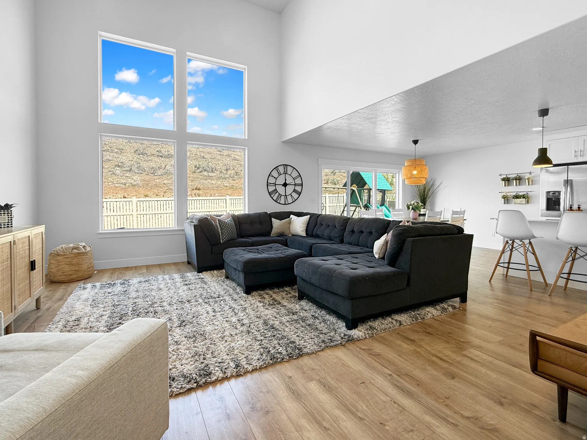 Open-concept living space featuring high ceilings, large picture windows, wood-finish flooring, and a kitchen area with pendant lighting and built-in shelving