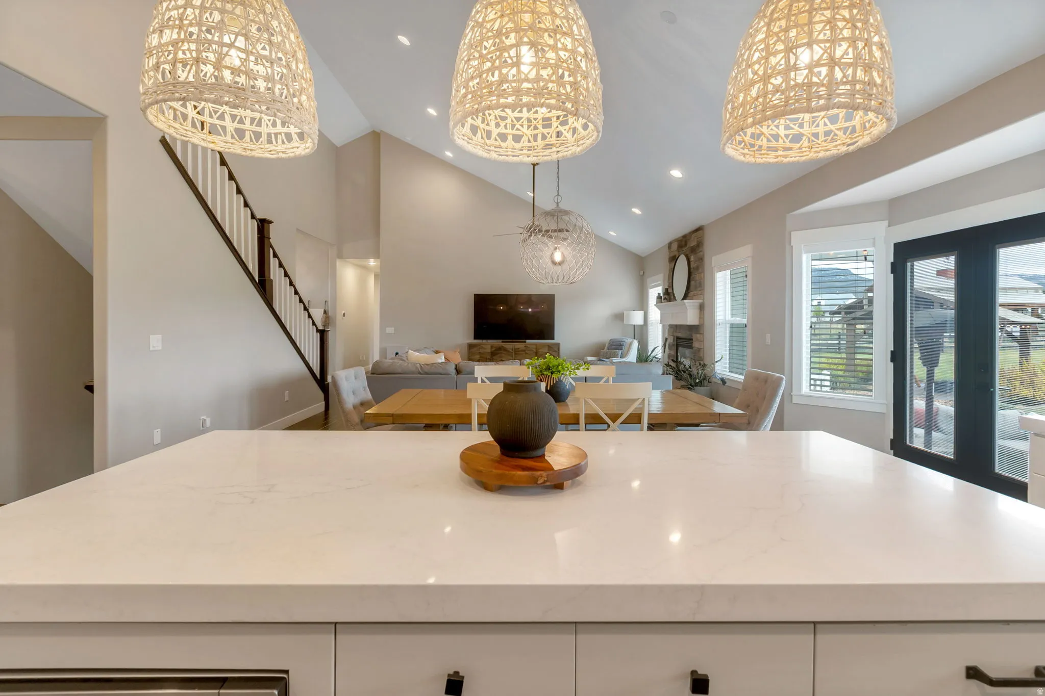 Kitchen island featuring a light-toned countertop, three woven pendant lights, vaulted ceilings, a stone fireplace, and double exterior doors