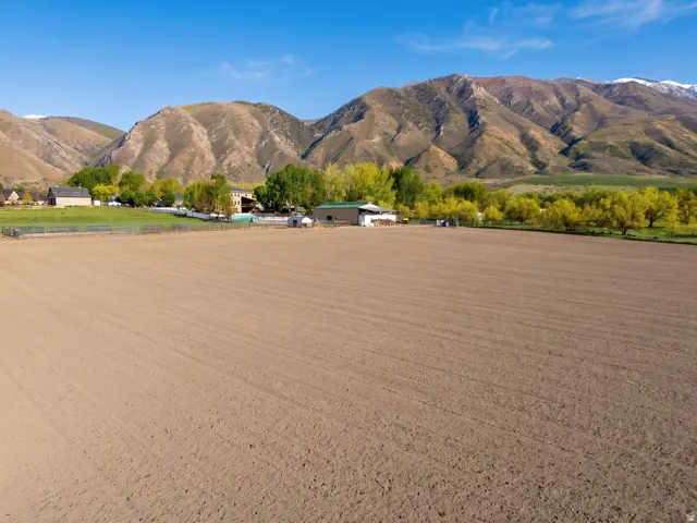 Expansive open field with tilled earth, bordered by mature trees and distant residential structures
