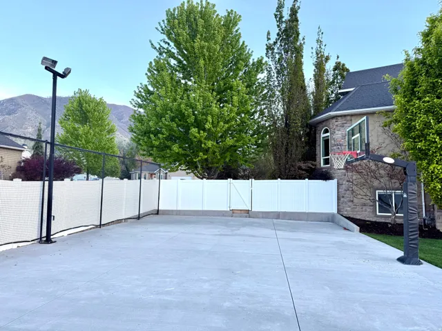 Paved sport court featuring a basketball hoop, perimeter fencing, and integrated lighting