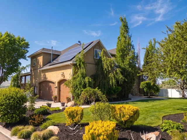 Stucco exterior residence featuring solar panels, arched garage doors, a stone pathway, and mature landscaping with manicured shrubs