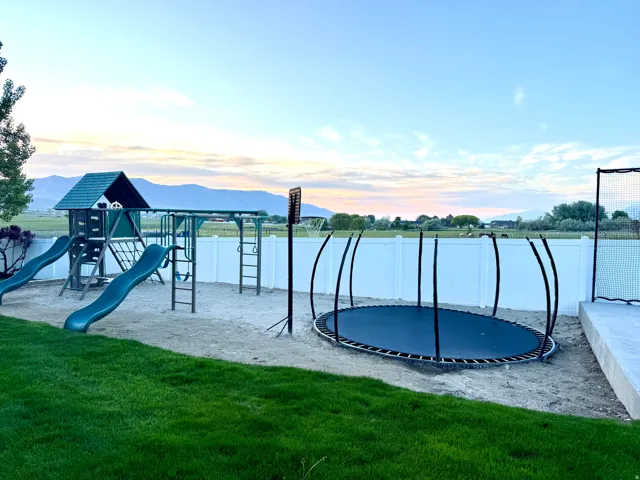 Expansive backyard featuring a manicured lawn, white vinyl fencing, a sand play area with a playset, and a built-in trampoline