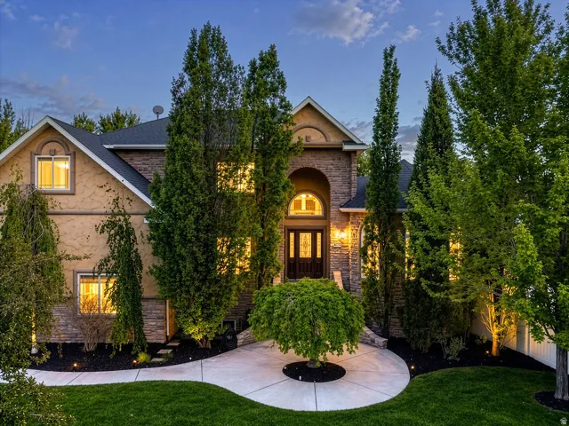 Stone and stucco exterior featuring a grand arched entry with a double-door entrance, extensive landscaping with mature trees, a curved concrete pathway, and exterior lighting fixtures