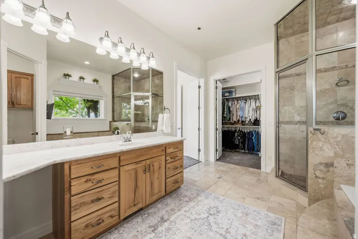 Primary Bathroom featuring a double vanity with wood cabinetry and a light-toned countertop, a large mirror with overhead vanity lighting, a fully enclosed shower with glass and stone-look tile, and natural light from a window