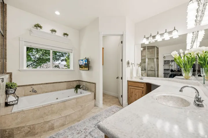 Primary Bathroom featuring a built-in jetted tub with tile surround, a spacious double vanity with integrated sinks, light-toned stone countertops, and a large wall-mounted mirror with vanity lighting