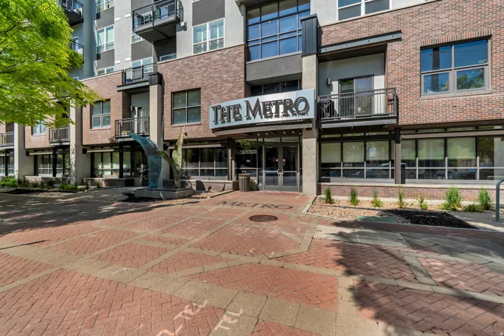 Modern multi-story building featuring brick and paneling exterior, ground-level entry with "THE METRO" signage, multiple balconies with metal railings, large storefront windows, and a paver walkway with a prominent art installation