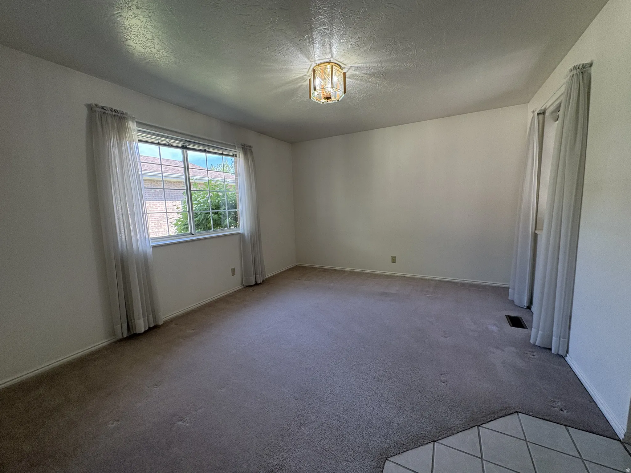 Spacious room featuring neutral carpeting, a large window with natural lighting, textured ceiling, and a decorative ceiling light fixture