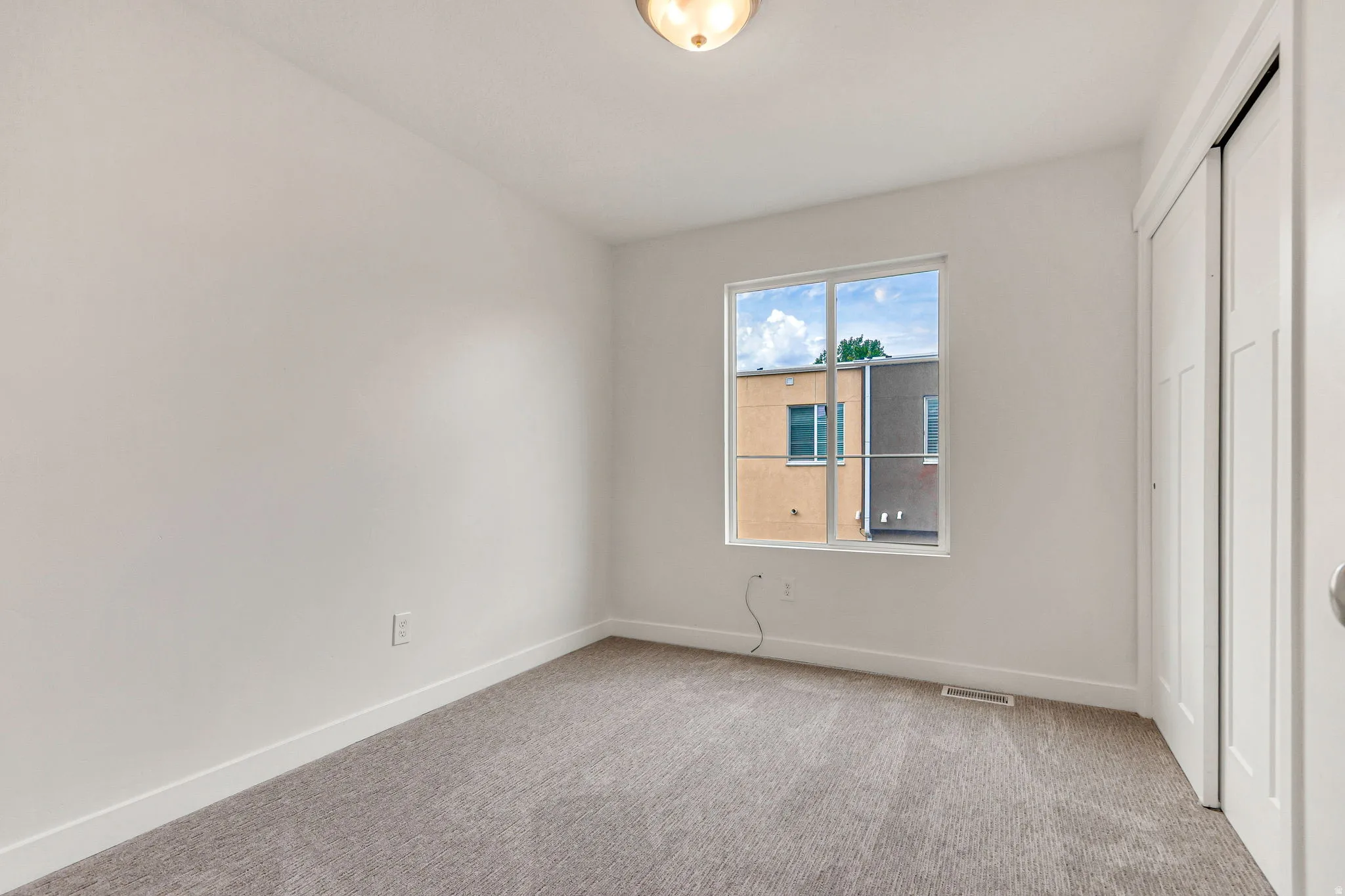 Bright room featuring a large window, plush gray carpeting, white baseboards, and a built-in closet with bi-fold doors