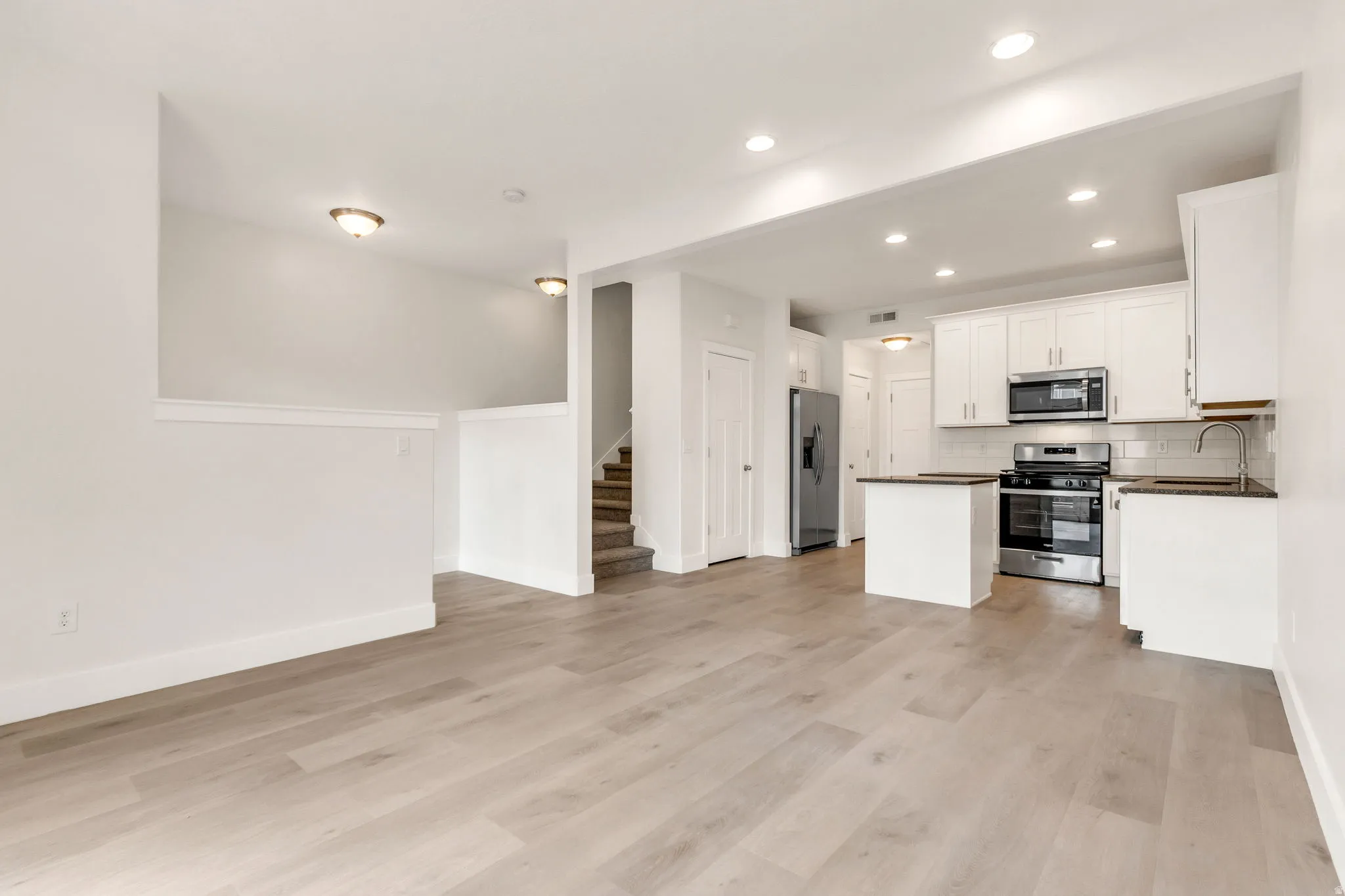 Open-concept living area featuring wood-finish flooring, recessed lighting, and a kitchen with white cabinetry, stainless steel appliances, and a tiled backsplash