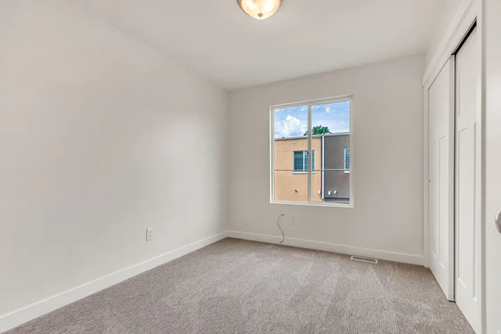 Bright room featuring a large window, plush gray carpeting, white baseboards, and a built-in closet with bi-fold doors