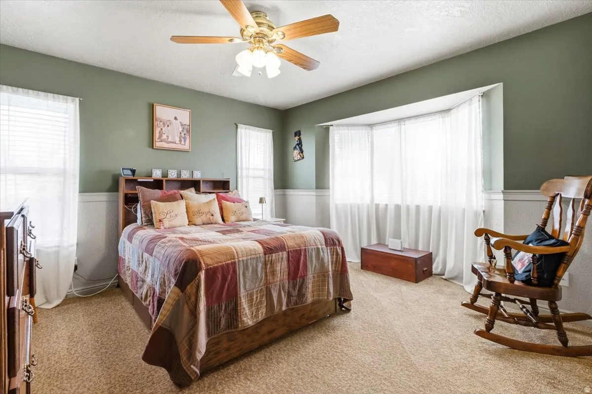 Spacious room featuring a bay window, ceiling fan with light fixture, carpeted flooring, and wainscoting