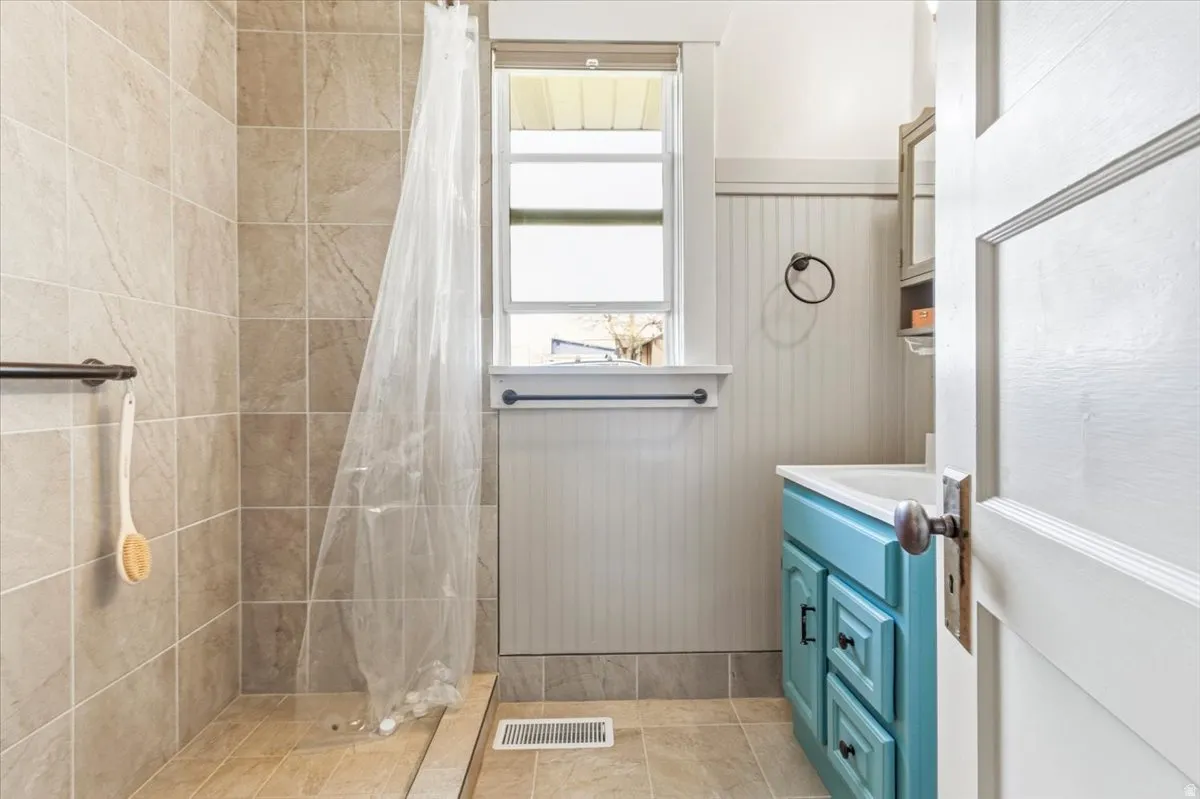 Bathroom featuring a tiled shower stall with a grab bar, a window with a privacy blind, beadboard wainscoting, and a blue vanity with a white basin sink