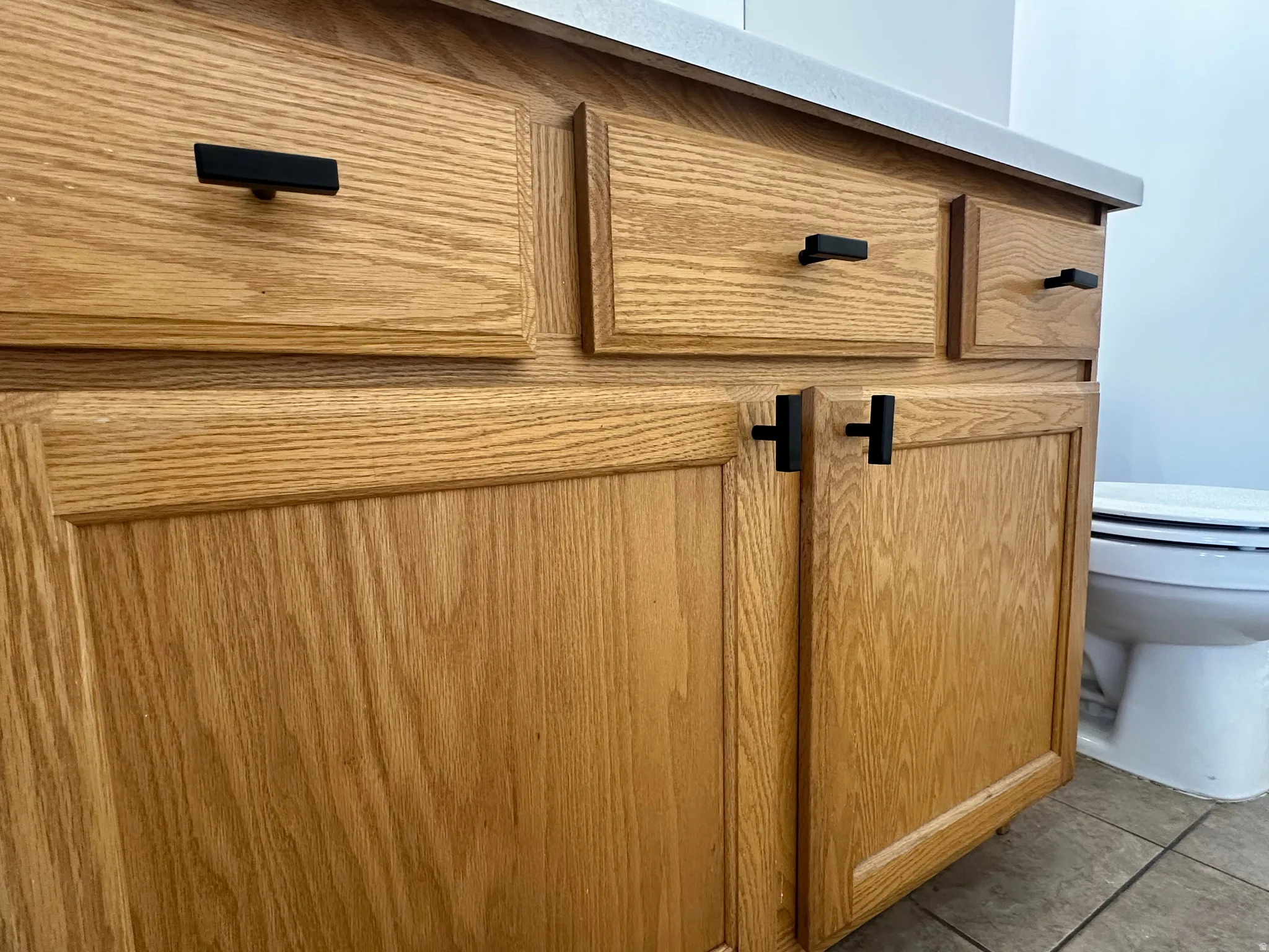 Oak wood-finish cabinetry with black hardware, a light-toned countertop, and tiled flooring
