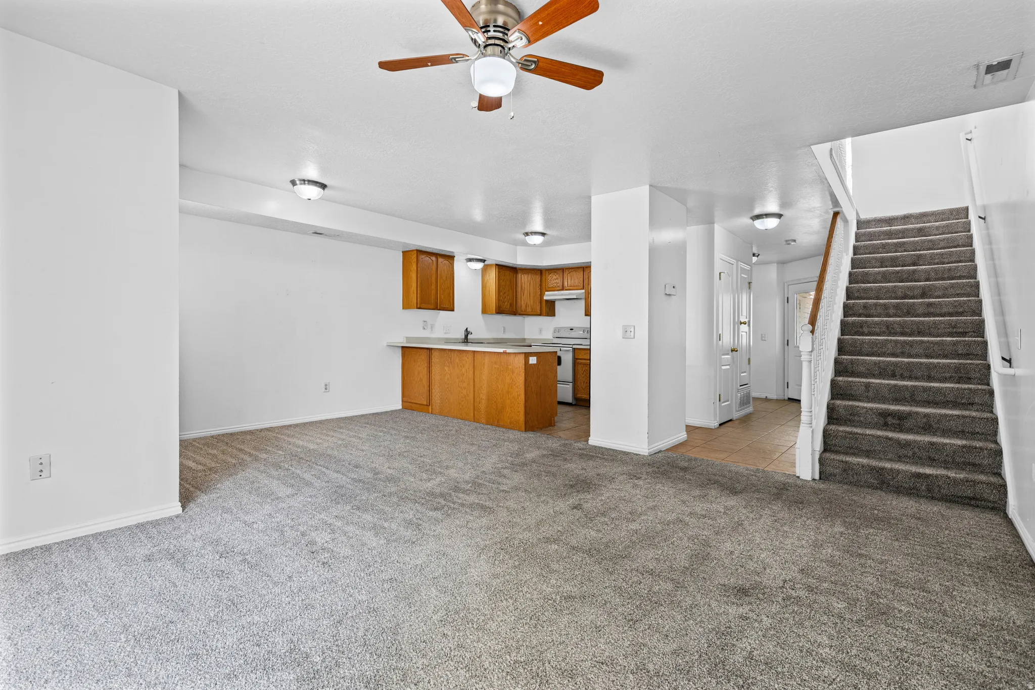 Open-concept living space featuring gray carpeting, a kitchen area with wood cabinetry, and a staircase with a carpeted runner