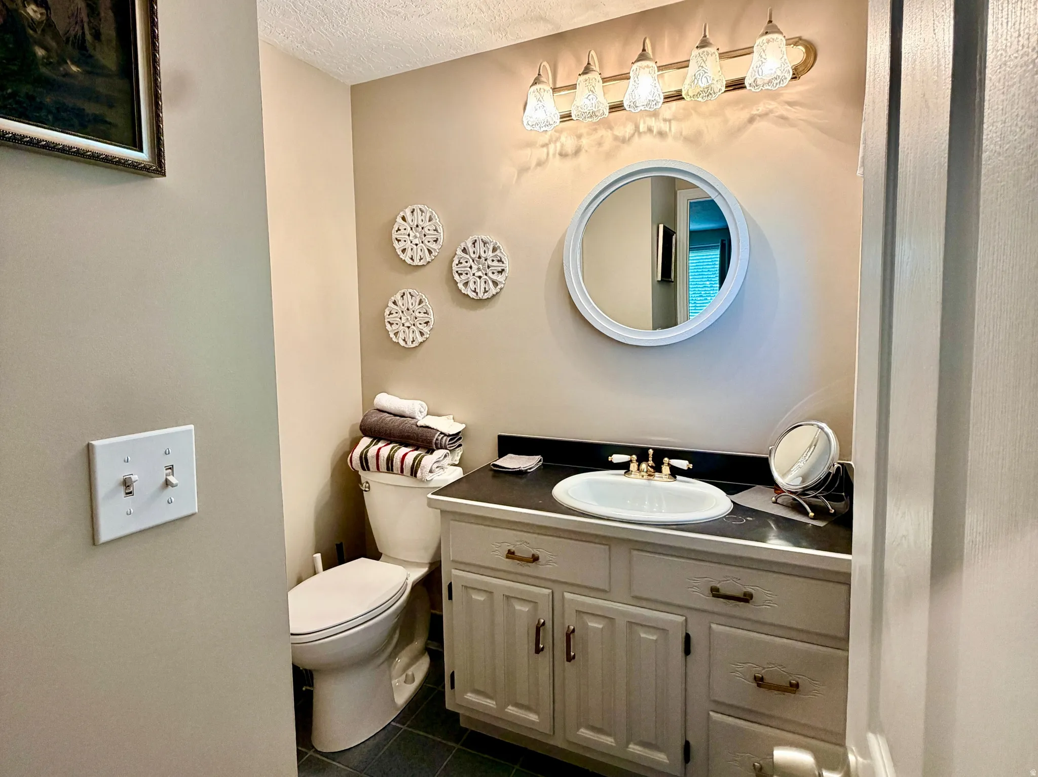 Bathroom featuring a light-colored vanity with a white basin and dark countertop, complemented by a round mirror and a five-light fixture