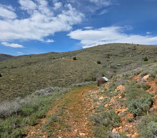 Expansive rural landscape featuring a single-track path, varied low-lying vegetation, distant hills, and a small rustic structure