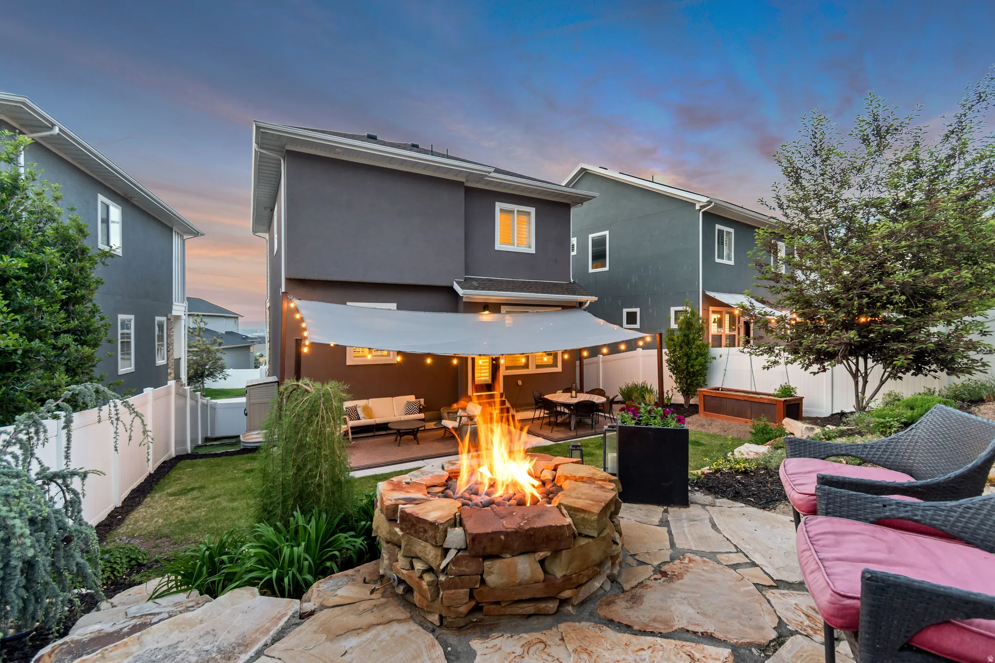 Expansive backyard featuring a stone-clad fire pit, flagstone patio, covered outdoor living area, landscaped garden beds, and perimeter fencing