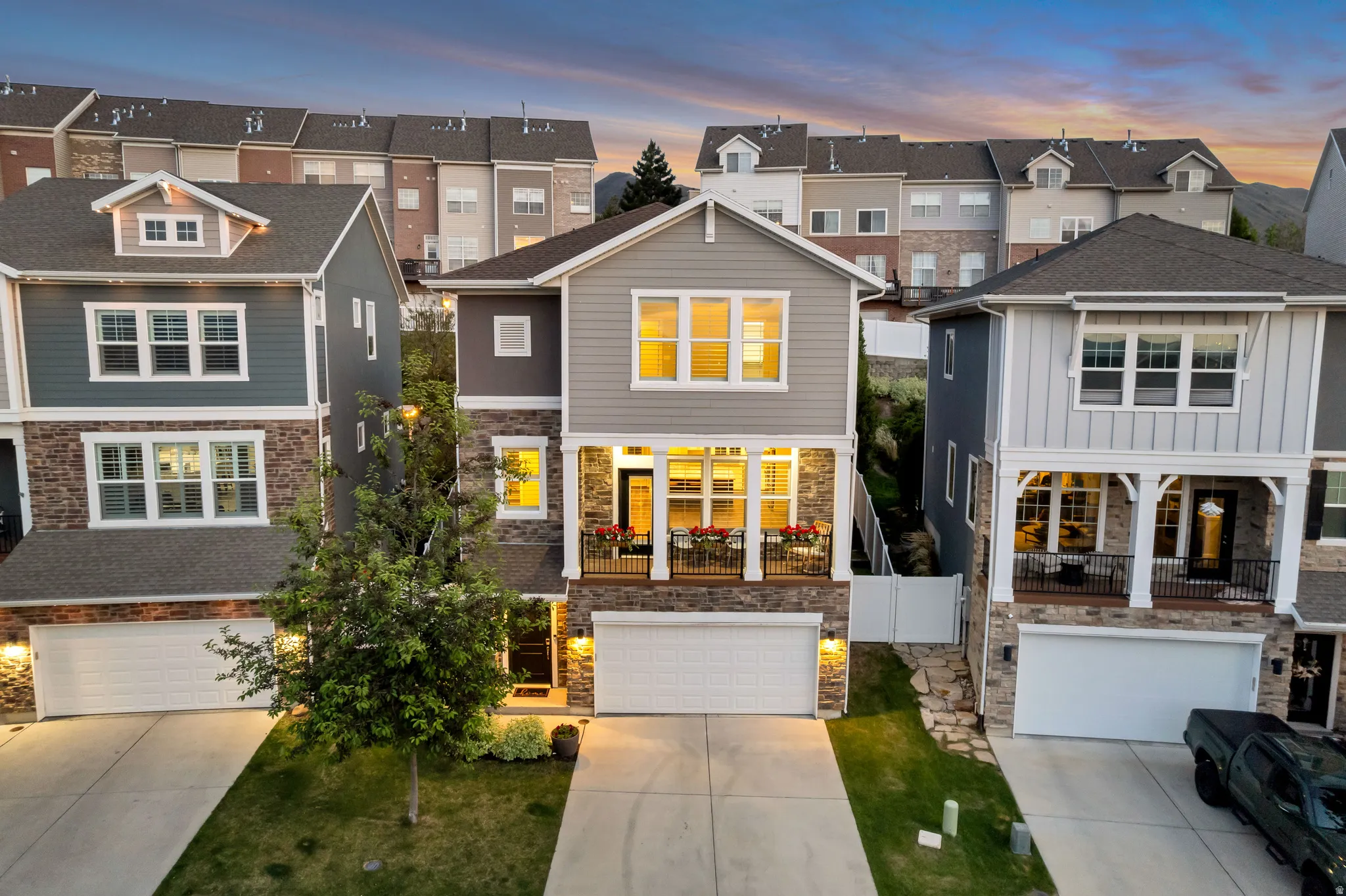 Contemporary multi-story residence featuring a stone and siding exterior, an attached two-car garage, an upper-level balcony, a covered entry porch, and mature landscaping