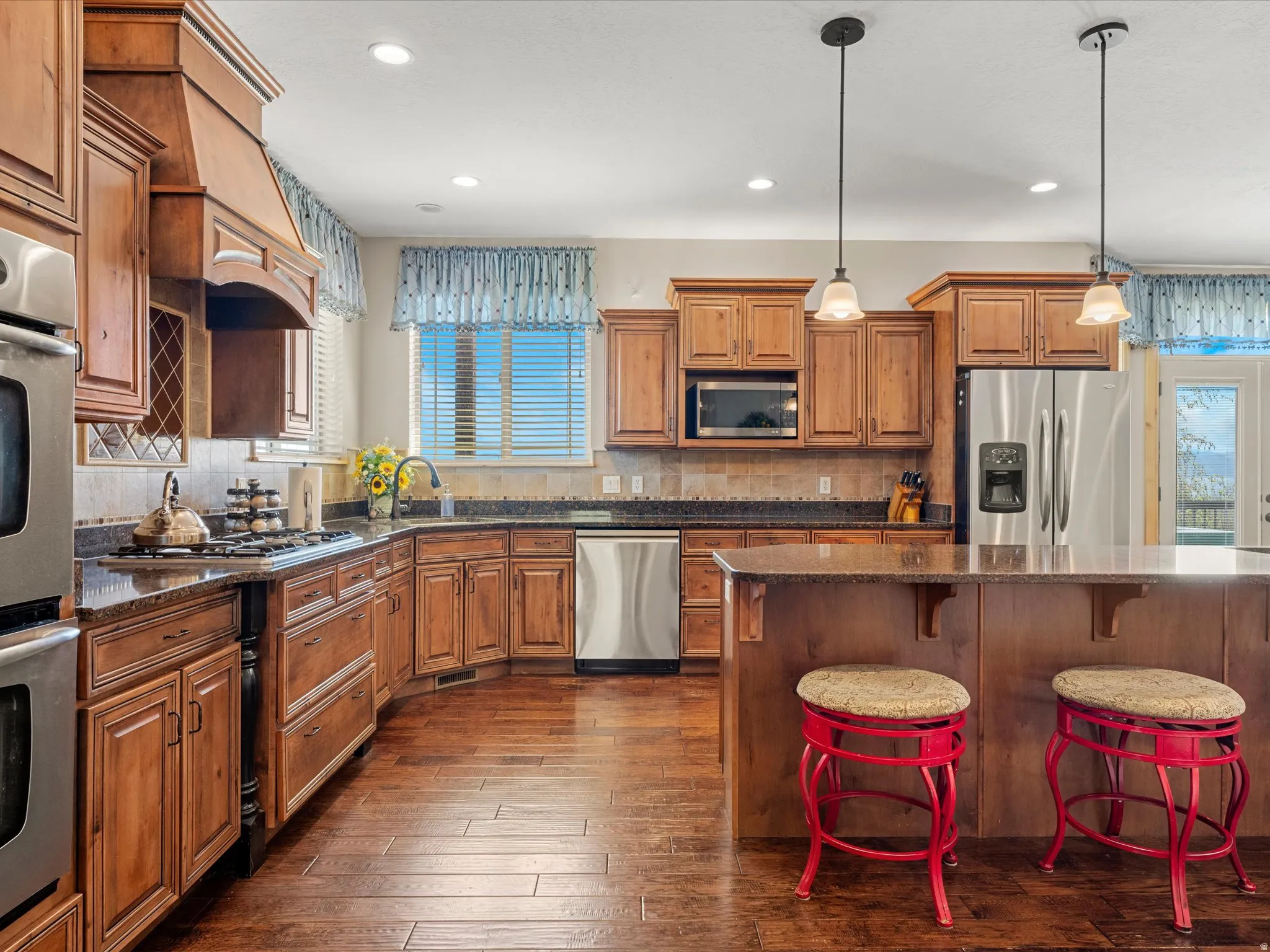 Kitchen featuring extensive wood cabinetry, dark countertops, stainless steel appliances, a center island with pendant lighting, and wood-finish flooring