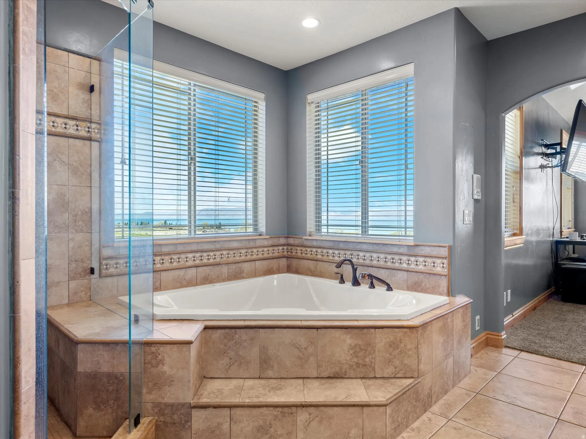 Corner soaking tub with tiled surround and integrated steps, dual windows with horizontal blinds, glass shower enclosure, and neutral-toned floor tiling