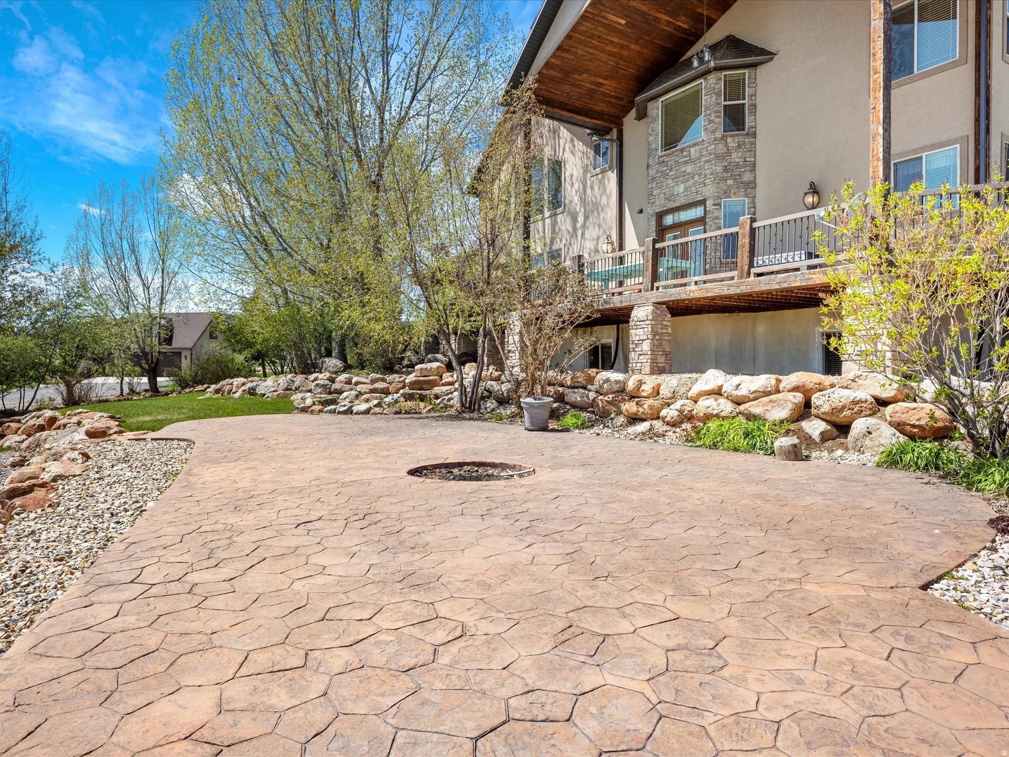 Stamped concrete patio with integrated fire pit, stone retaining walls, mature landscaping, and a multi-level deck with wood railing