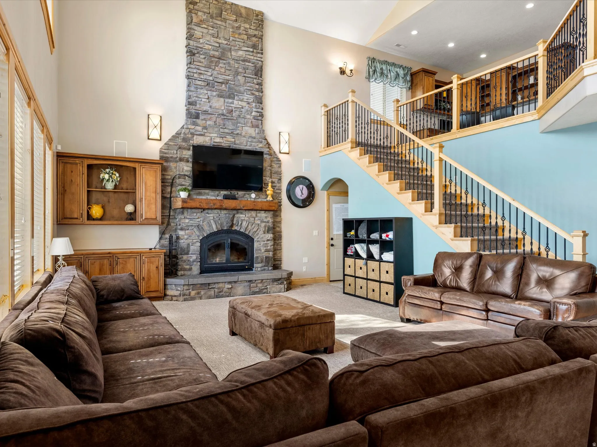 Two-story living area featuring a stone-clad fireplace with wood mantle, high ceilings, multiple windows with blinds, and a prominent staircase with wood railings and metal balusters