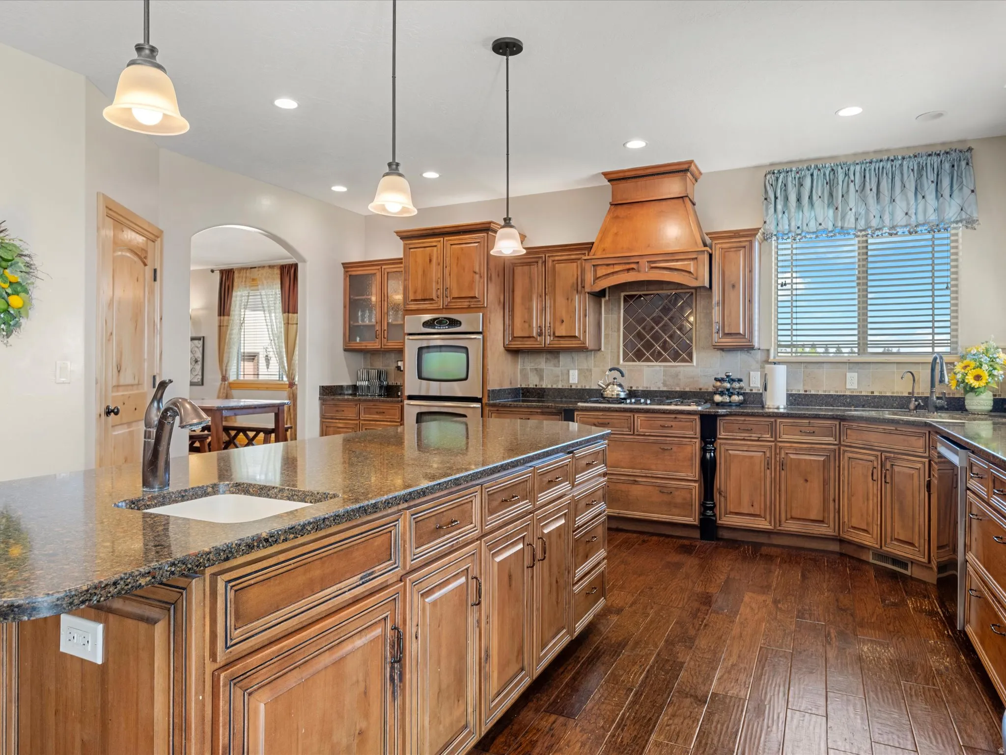 Spacious kitchen featuring a large central island with an undermount sink, rich wood-finish flooring, custom wood cabinetry, granite countertops, and stainless steel built-in double ovens
