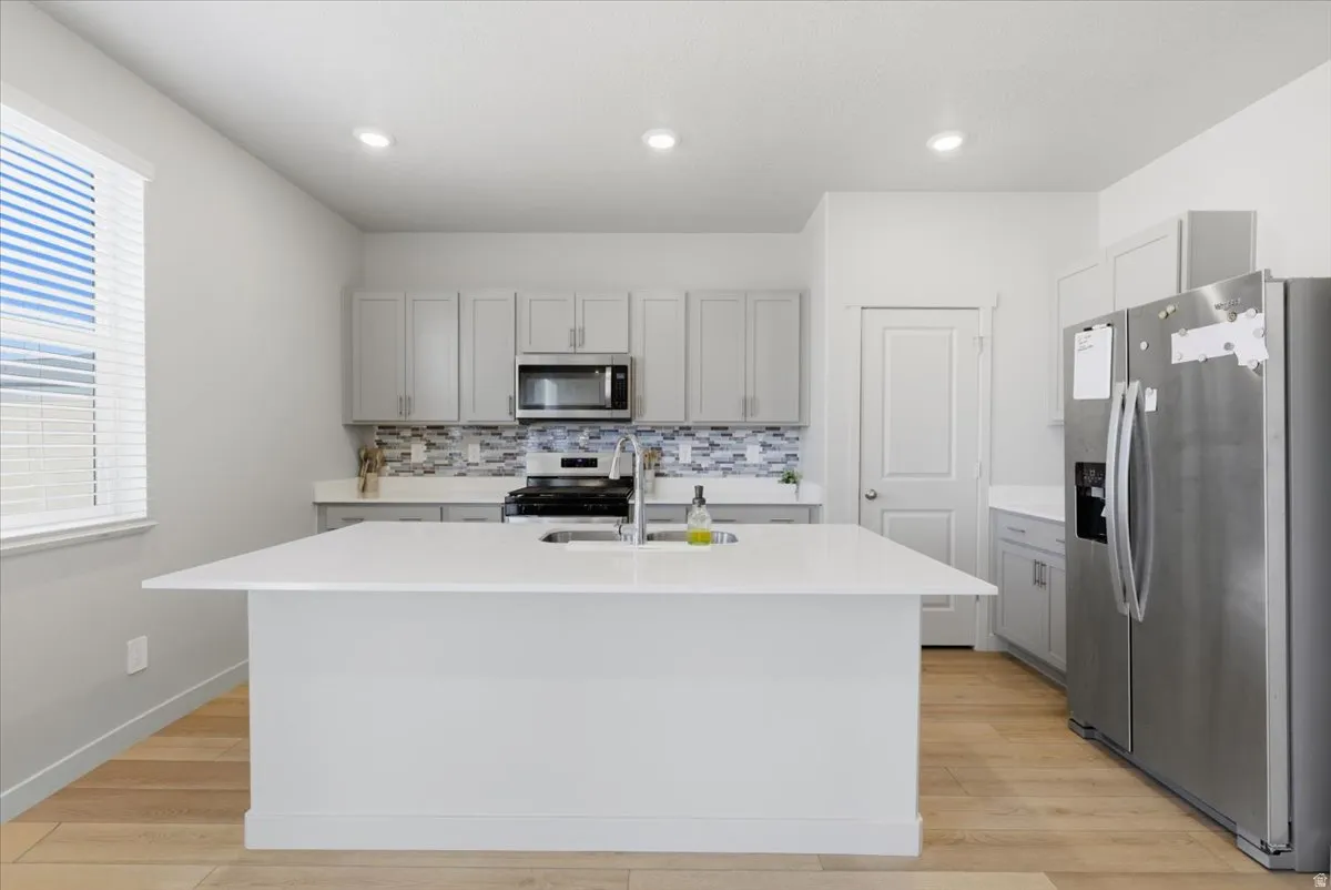 Modern kitchen featuring a large central island with an undermount sink, light wood-finish flooring, recessed lighting, stainless steel appliances, and sleek grey cabinetry