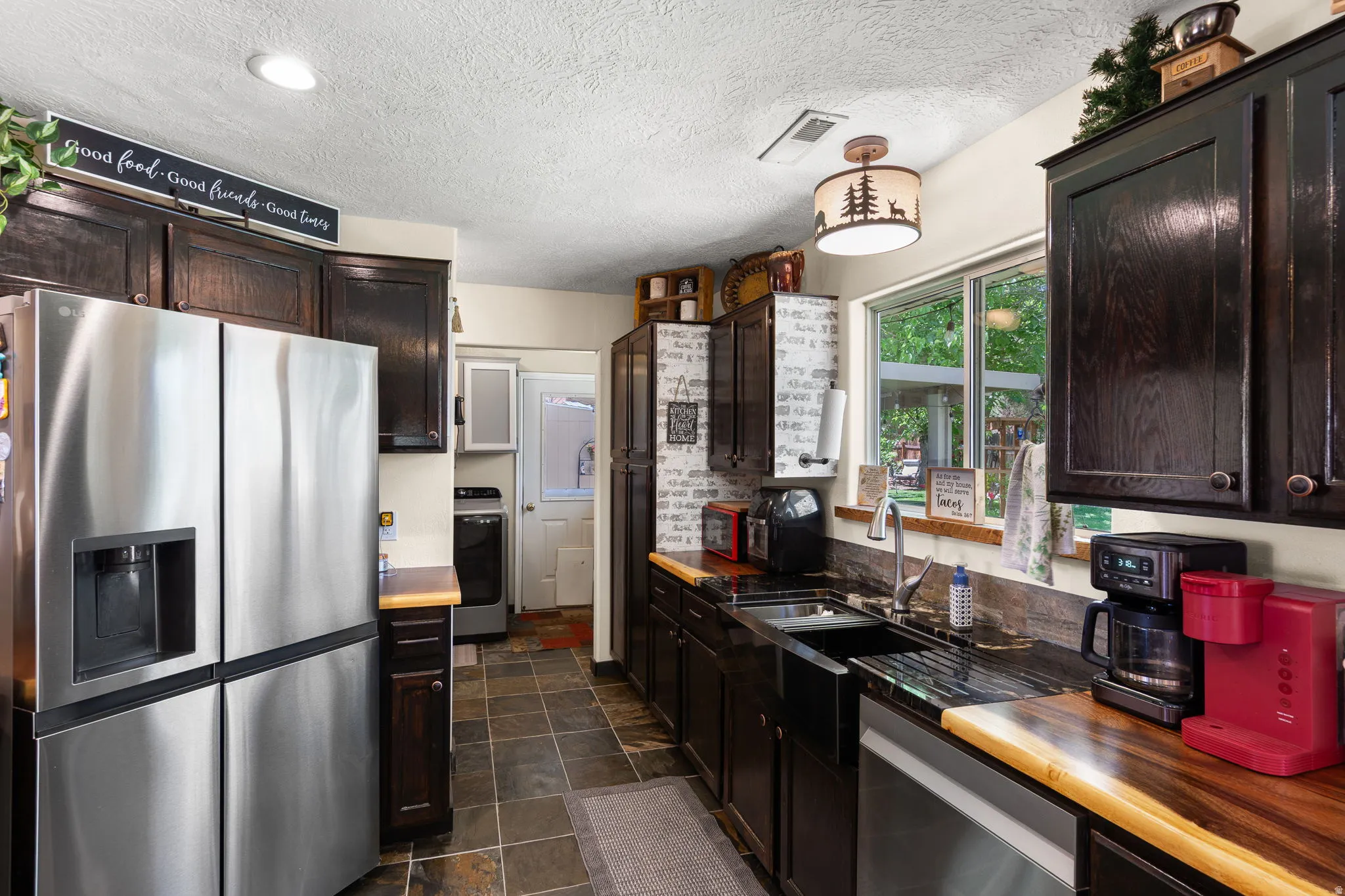 Kitchen featuring rich dark wood cabinetry, a stainless steel refrigerator, stone tile flooring, a black farmhouse-style sink, and a window with outdoor landscaping views