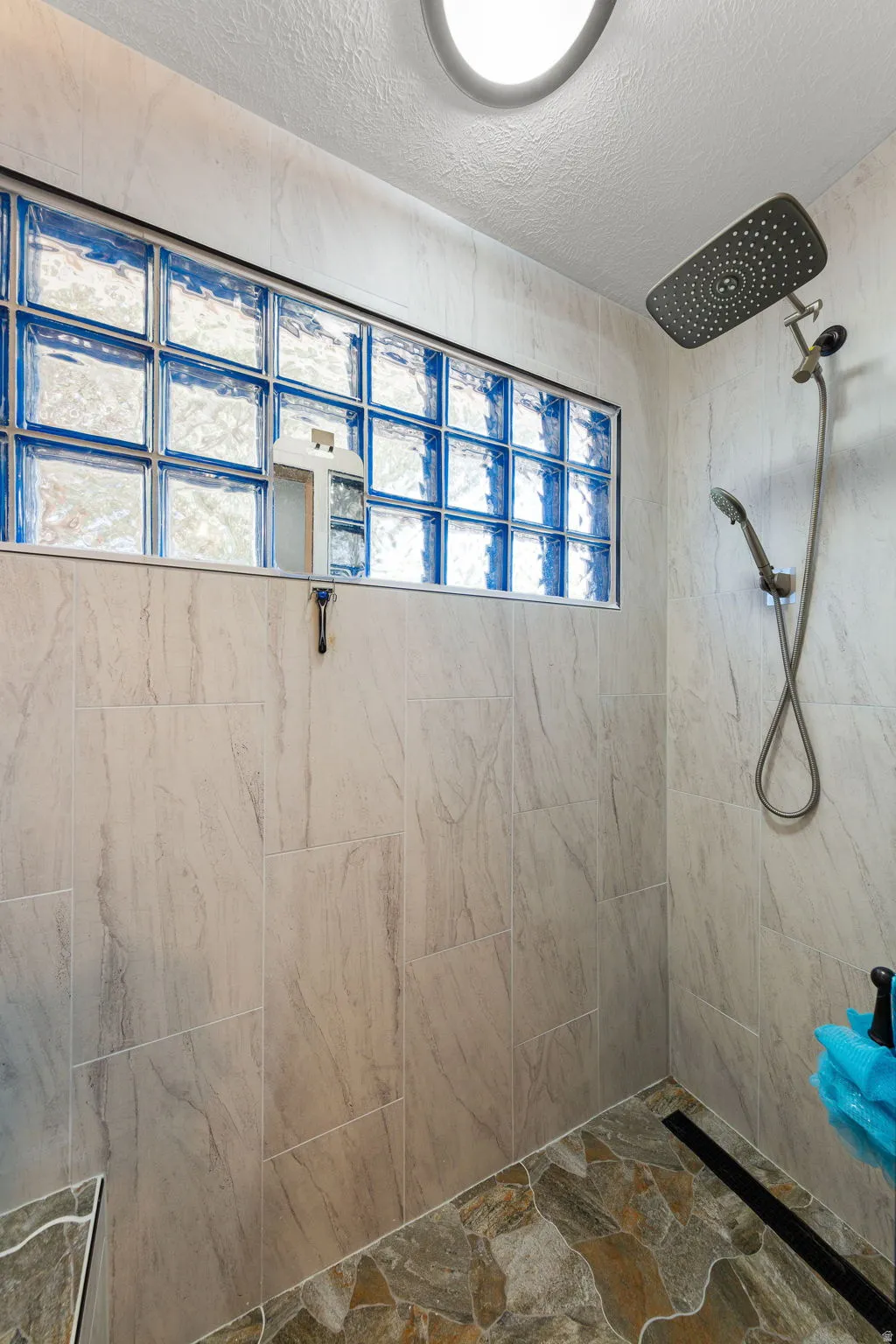 Walk-in shower featuring a rain shower head, handheld shower, full-height wall tiling, glass block window, and flagstone-style flooring with a linear drain