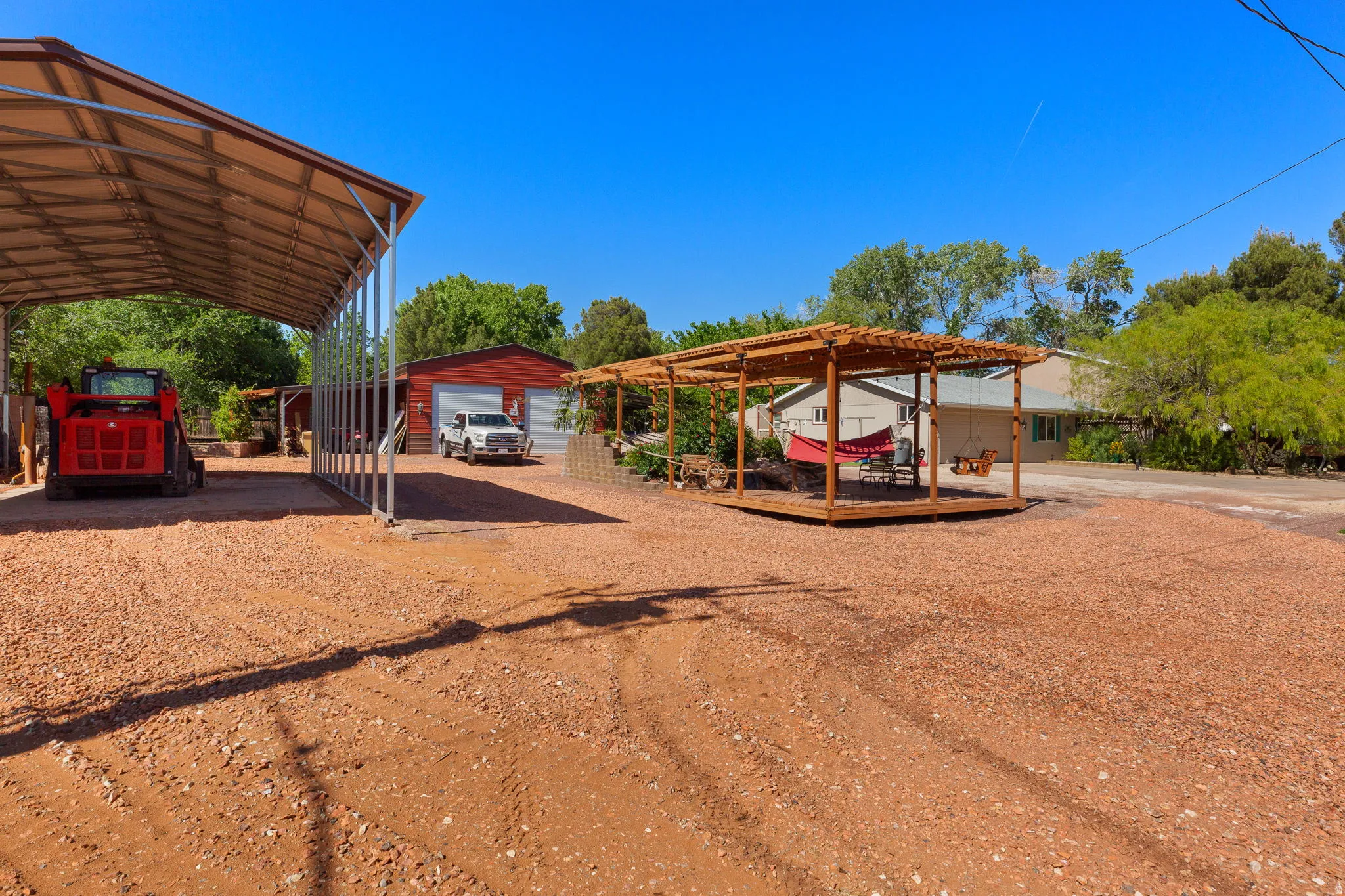 Expansive gravel lot featuring a large metal carport, a red metal outbuilding with two garage doors, and a wood-framed pergola with string lights