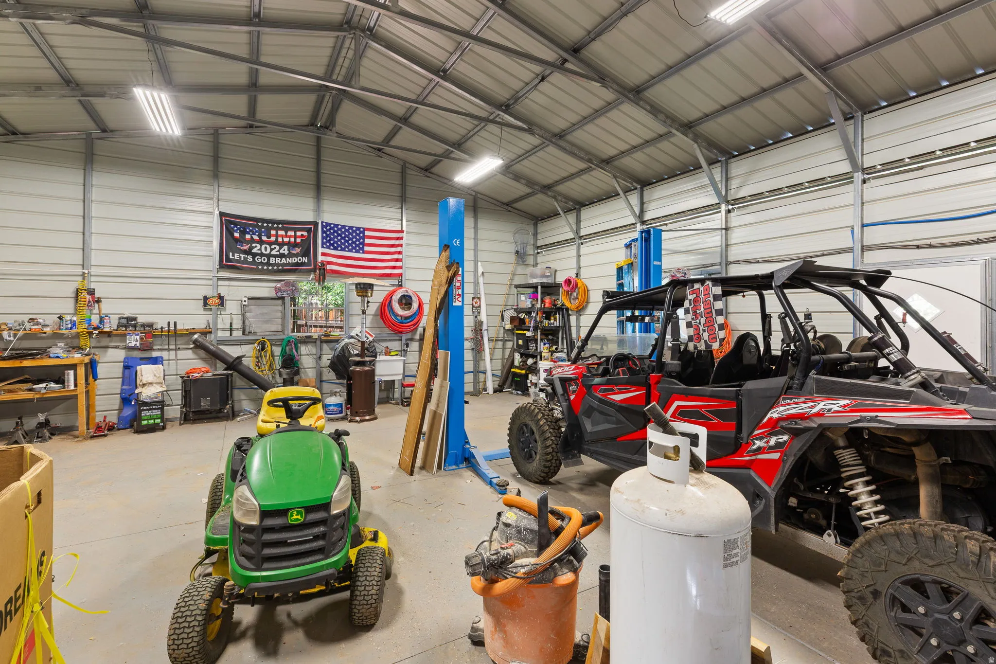 Spacious metal workshop featuring a concrete floor, exposed metal beam ceiling, fluorescent lighting, and a built-in vehicle lift
