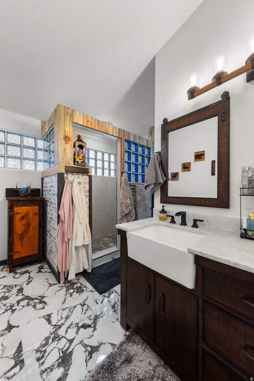 Bathroom featuring a white marble-look floor, a dark wood-finish vanity with a farmhouse sink, and a framed mirror with a three-bulb light fixture