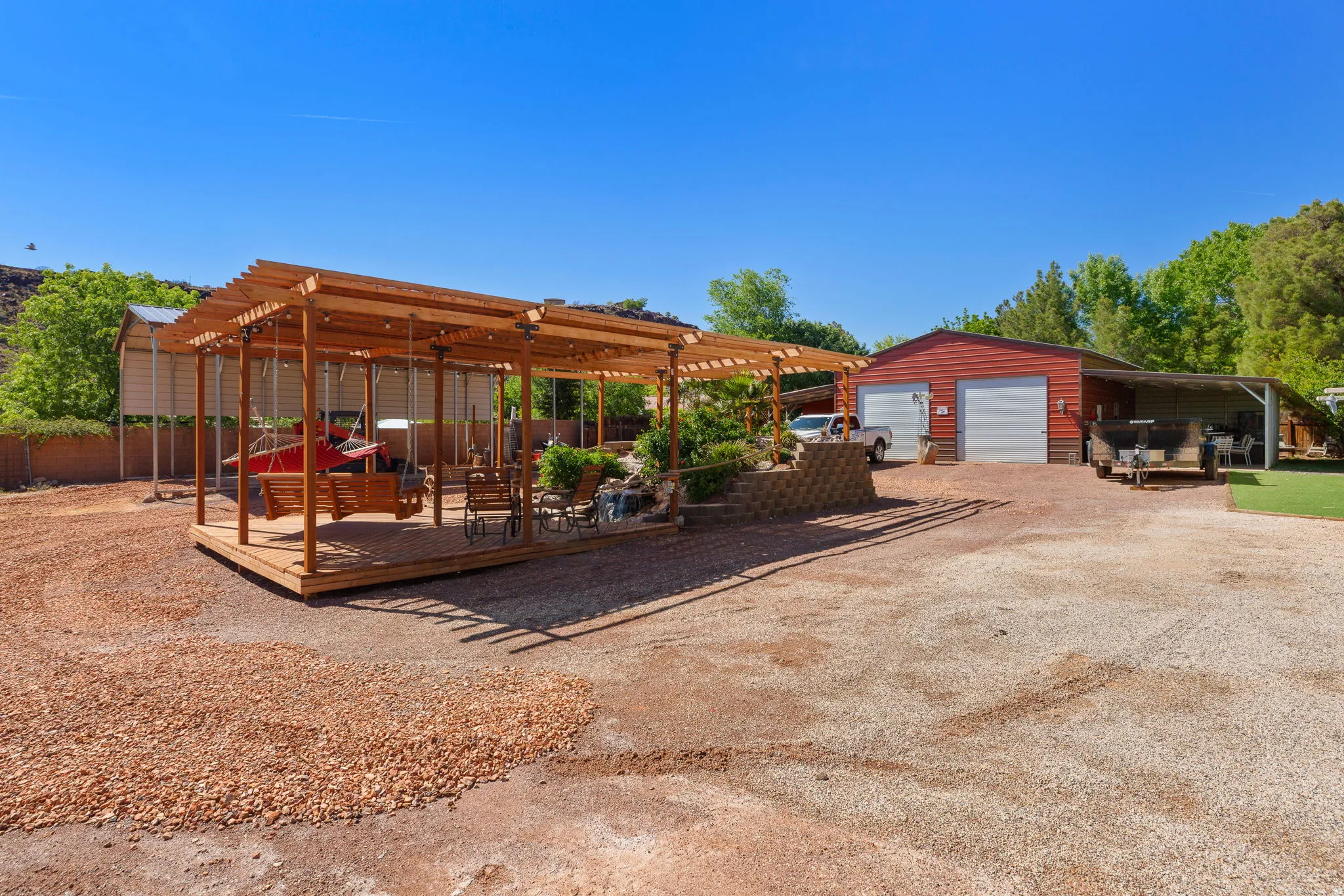Expansive gravel yard featuring a wood pergola-covered patio with string lighting, a large red two-bay garage, and an attached covered carport