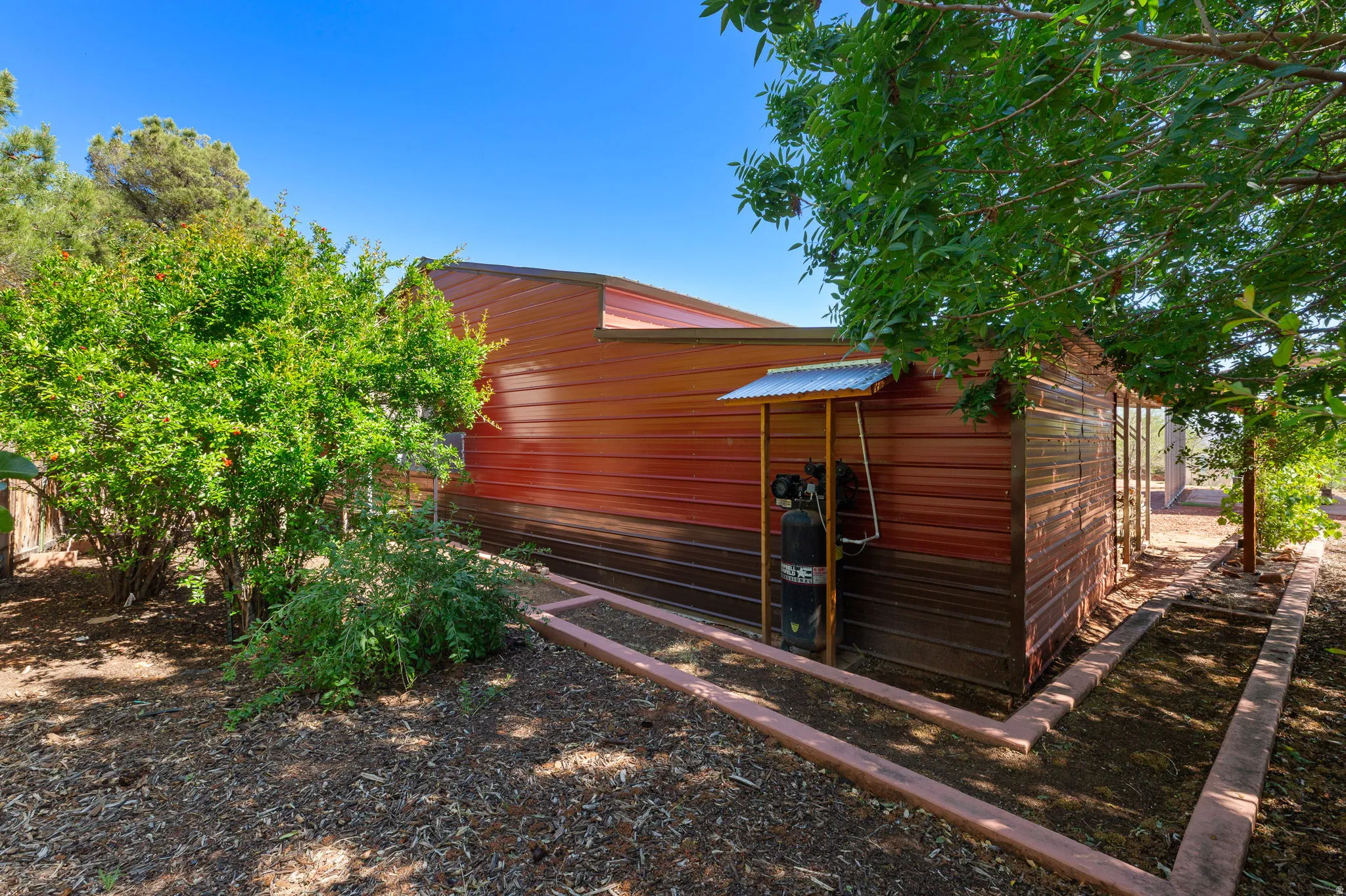 Metal-sided outbuilding featuring a two-tone brown and red exterior, a corrugated metal awning, and concrete garden bed borders