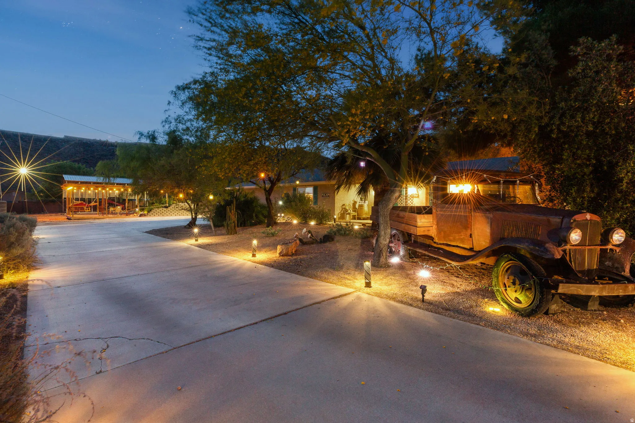 Spacious concrete driveway featuring integrated landscape lighting, mature trees, and a covered outdoor entertaining area