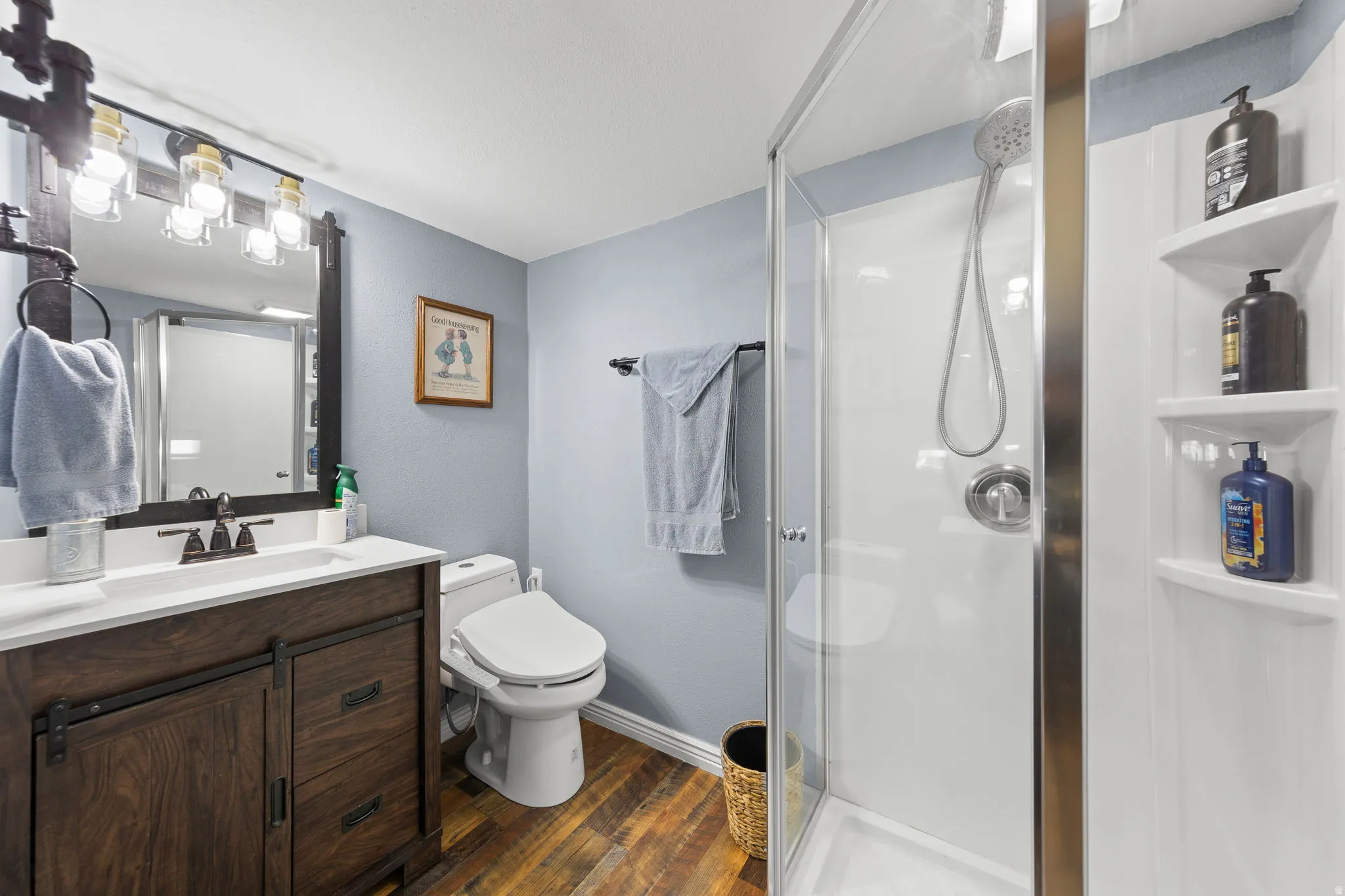 Bathroom featuring a clear glass shower enclosure, wood-finish flooring, a vanity with a white countertop and dark wood-look cabinetry, and an industrial-style light fixture above a dark-framed mirror