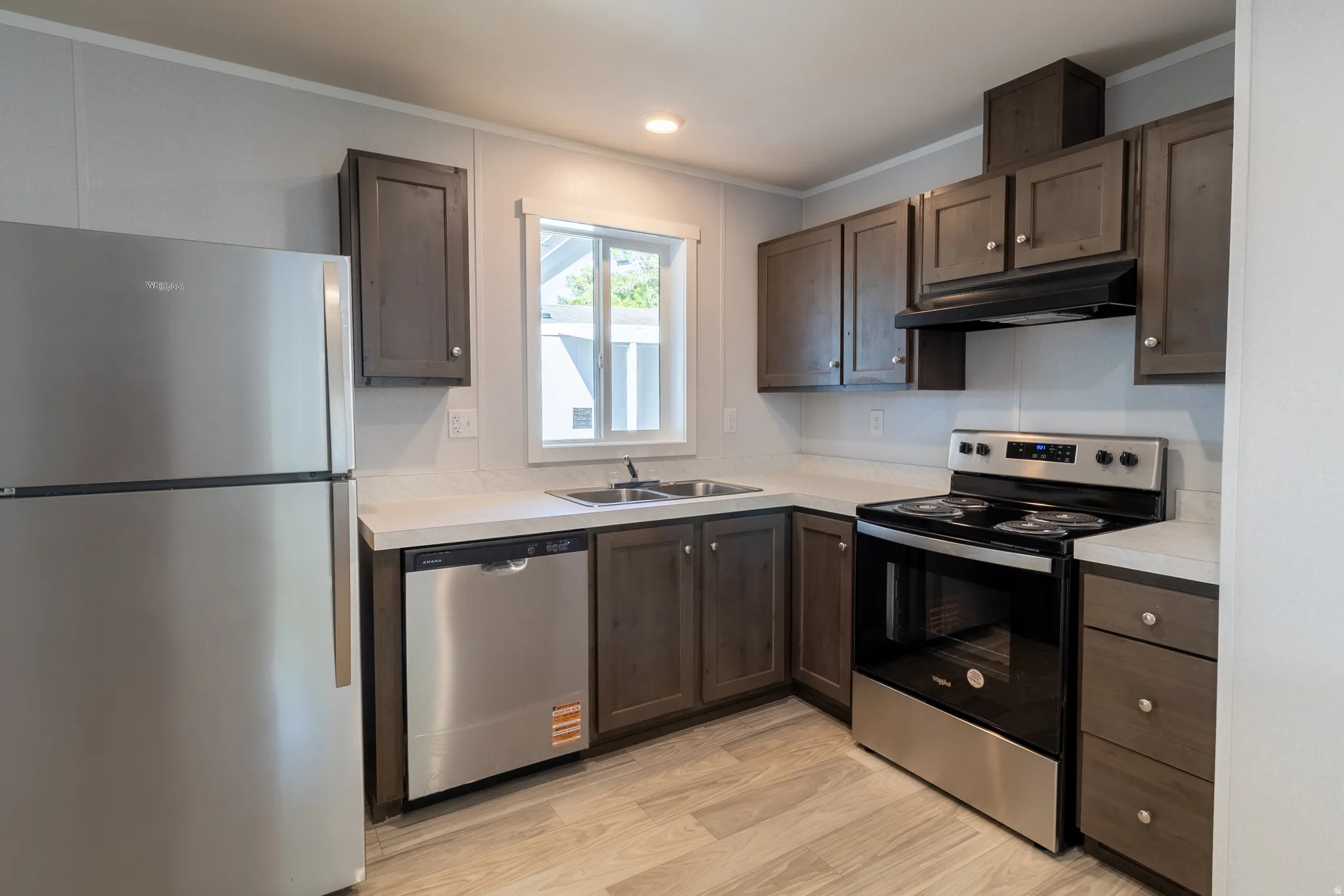 Kitchen with stainless steel appliances, wood-finish cabinetry, light countertops, and a window above the double-basin sink