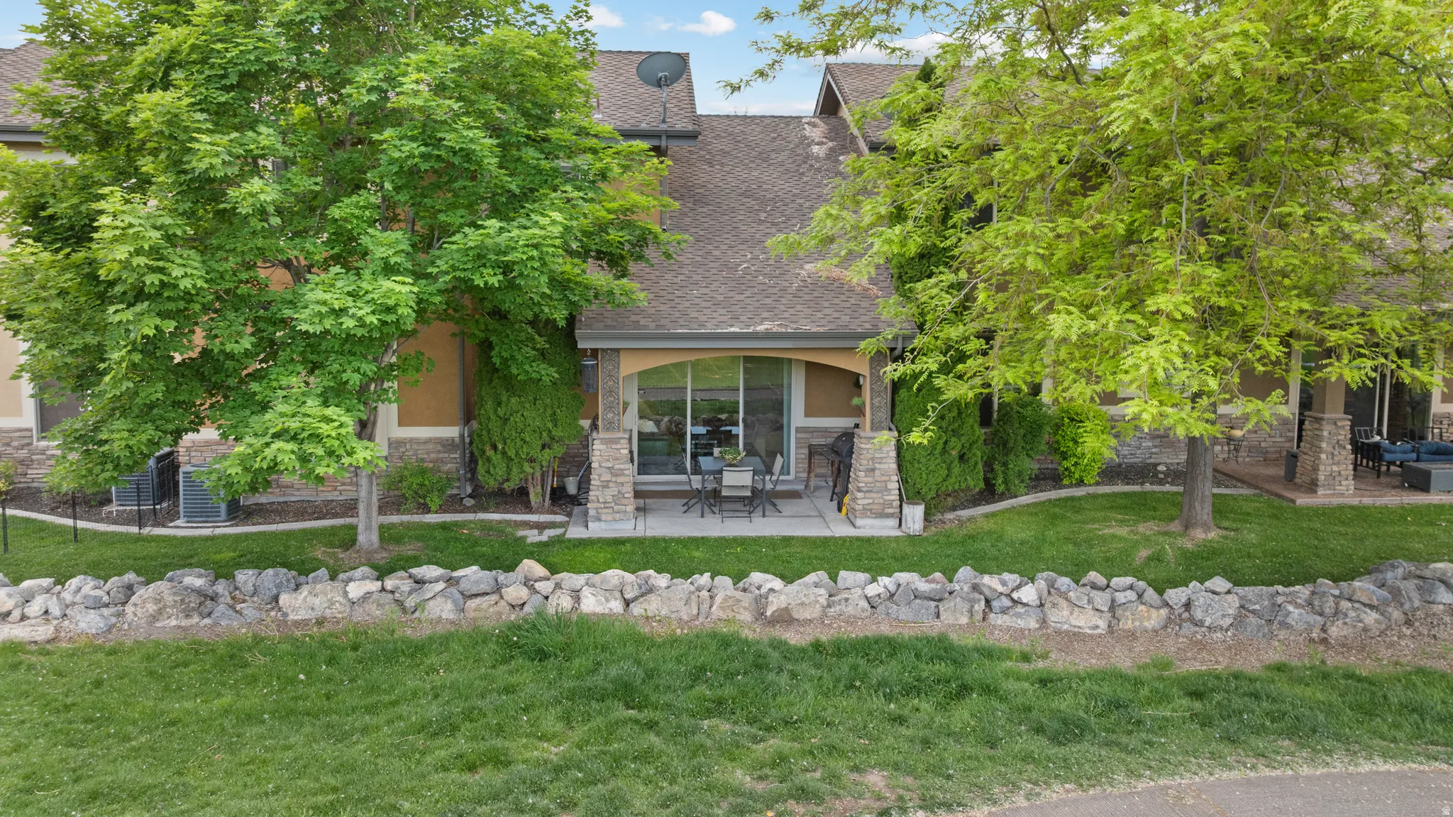Rear exterior featuring a covered patio with stone-wrapped columns, a sliding glass door, a manicured lawn, and mature trees