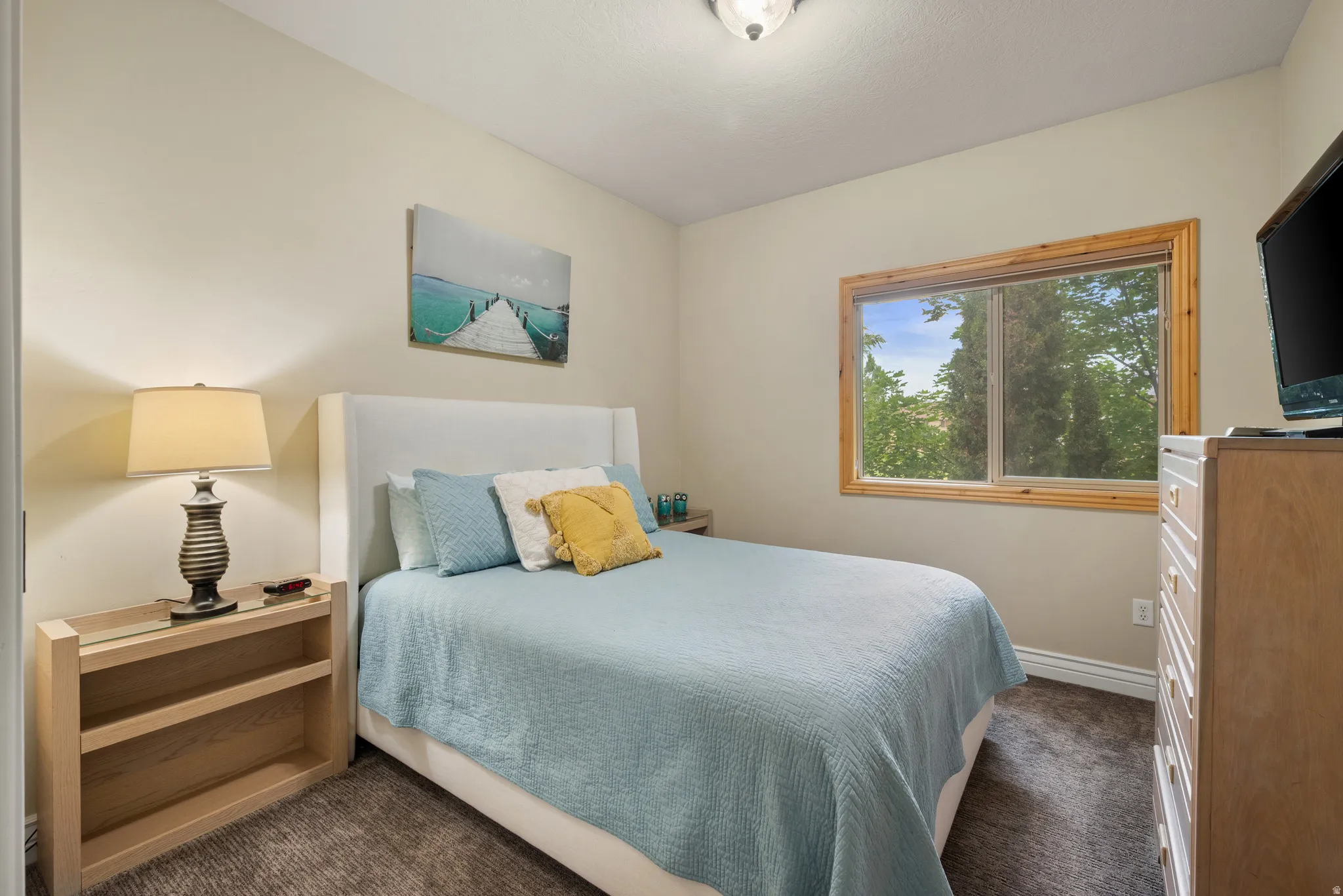 Well-lit room featuring light neutral wall paint, a window with natural wood trim, textured carpeting, and a ceiling-mounted light fixture