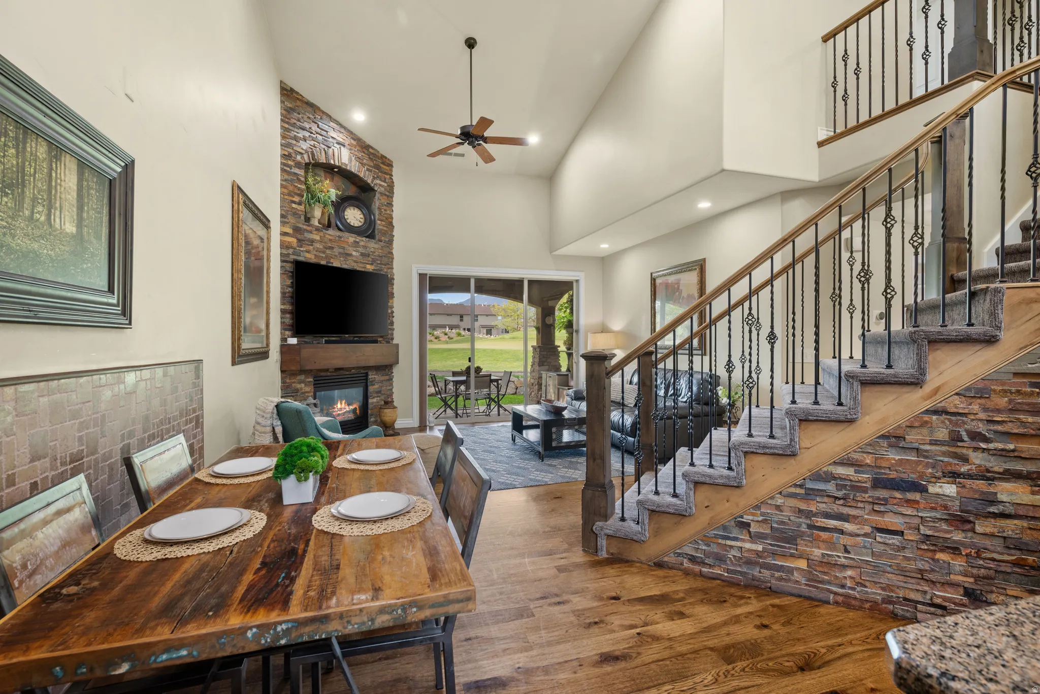 Open-concept interior featuring wood-finish flooring, a stone-clad fireplace, a staircase with decorative iron balusters, and a screened outdoor patio