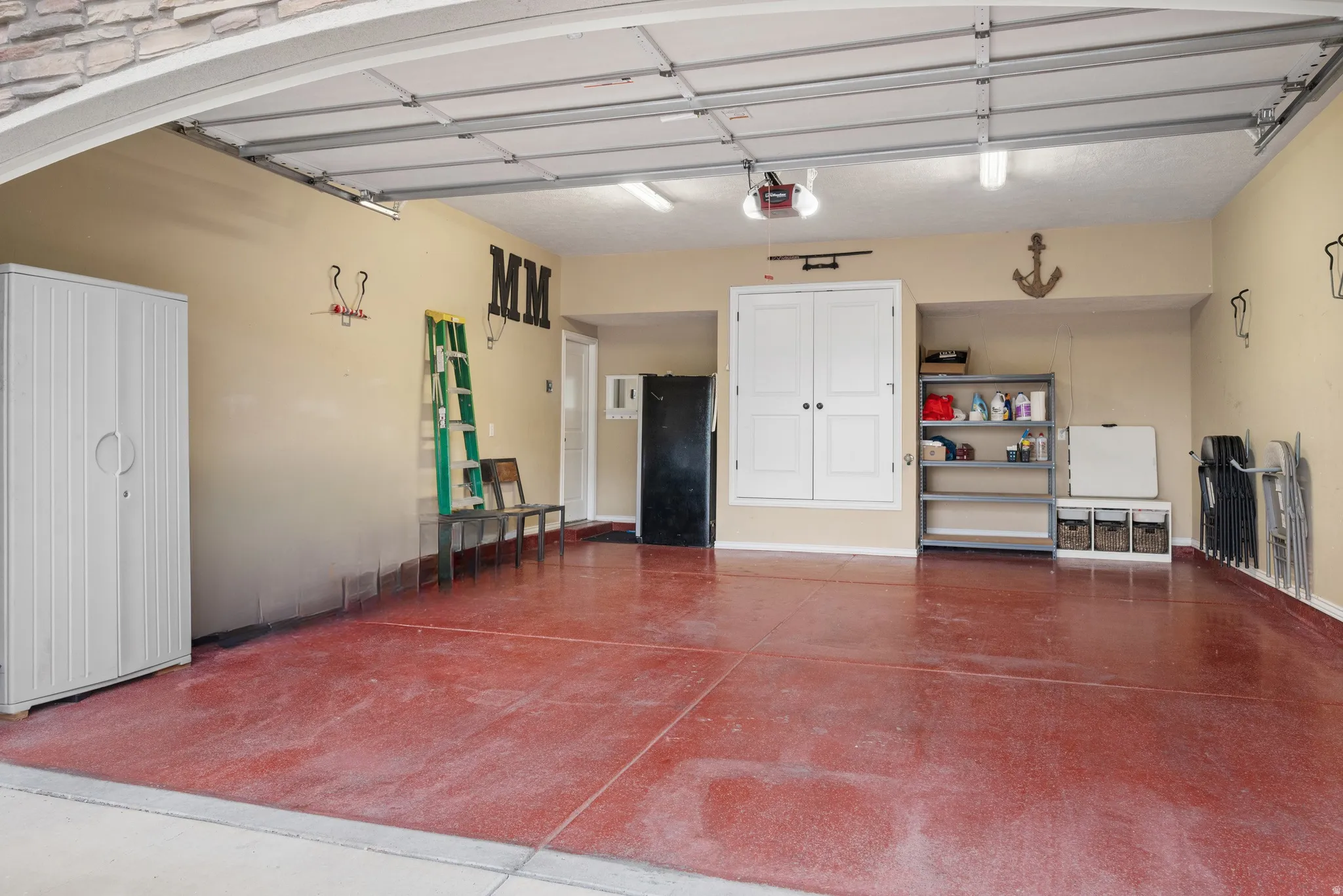 Spacious 2-car garage with a red-toned concrete floor, overhead lighting, and a built-in storage cabinet with white double doors