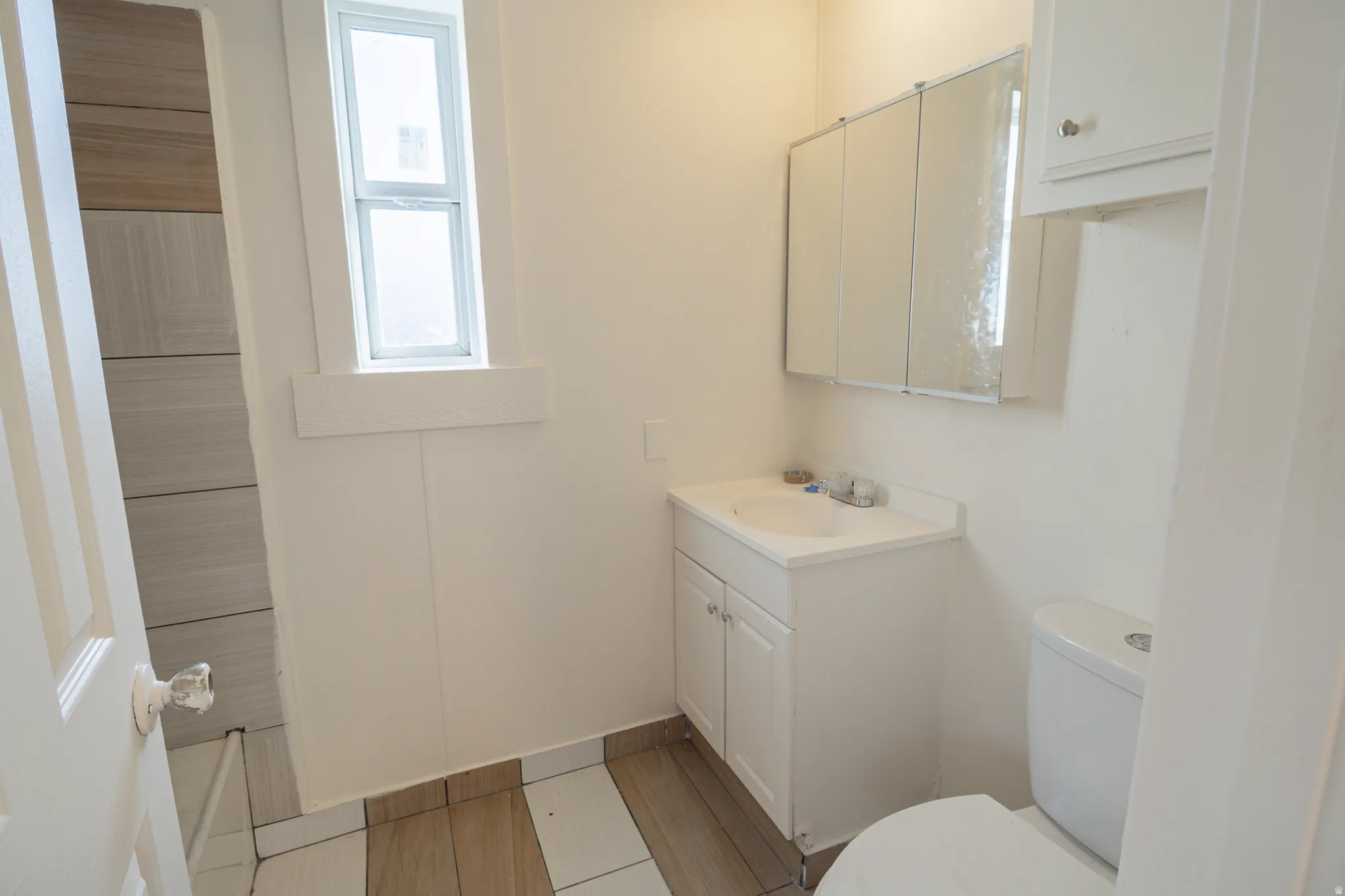 Full bathroom featuring a single window, white vanity with integrated sink, coordinating medicine cabinet, flush toilet, and wood-finish tile flooring