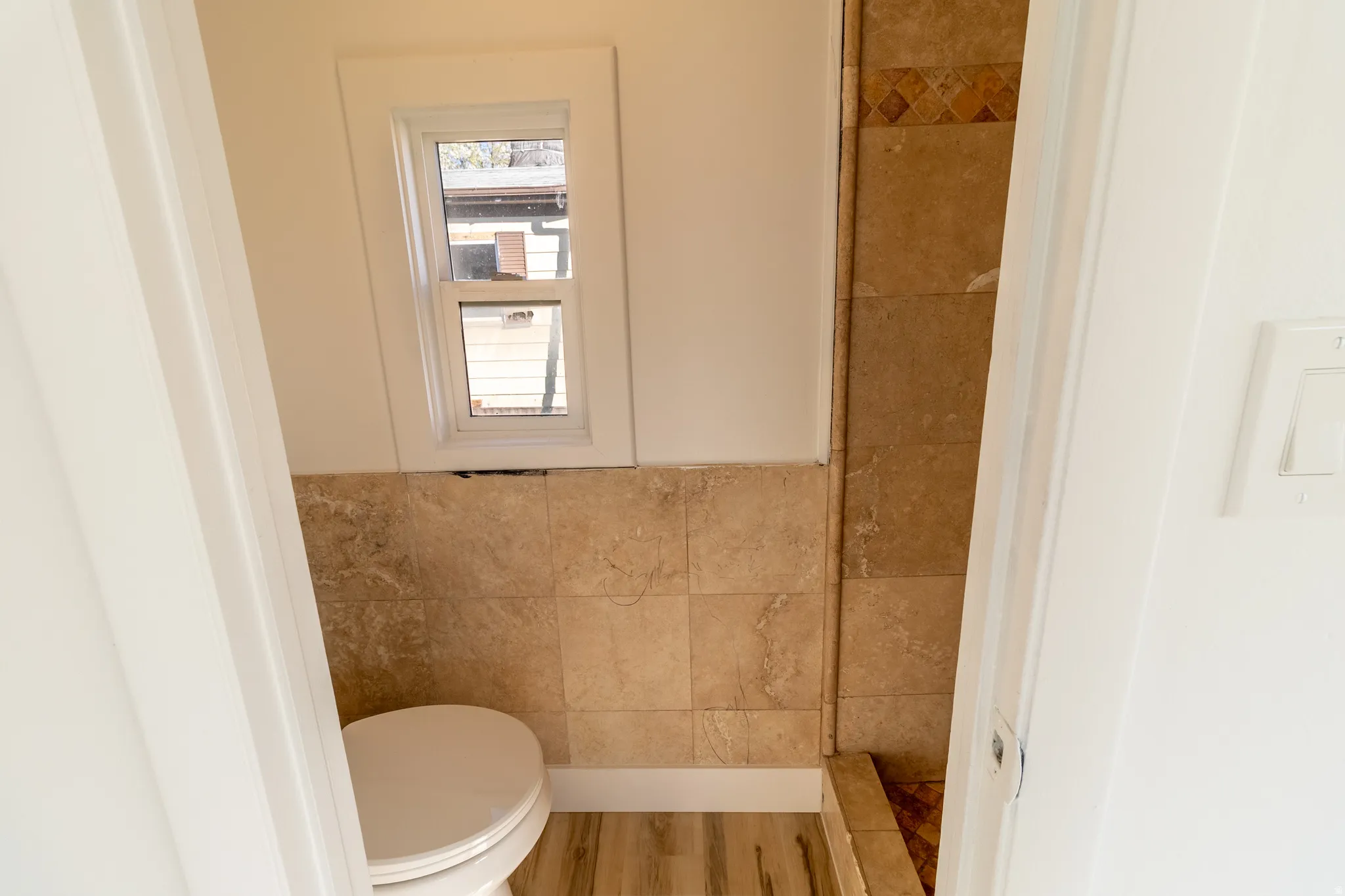 Compact bathroom featuring a white toilet, tan tile wainscoting, wood-finish flooring, and a window for natural light