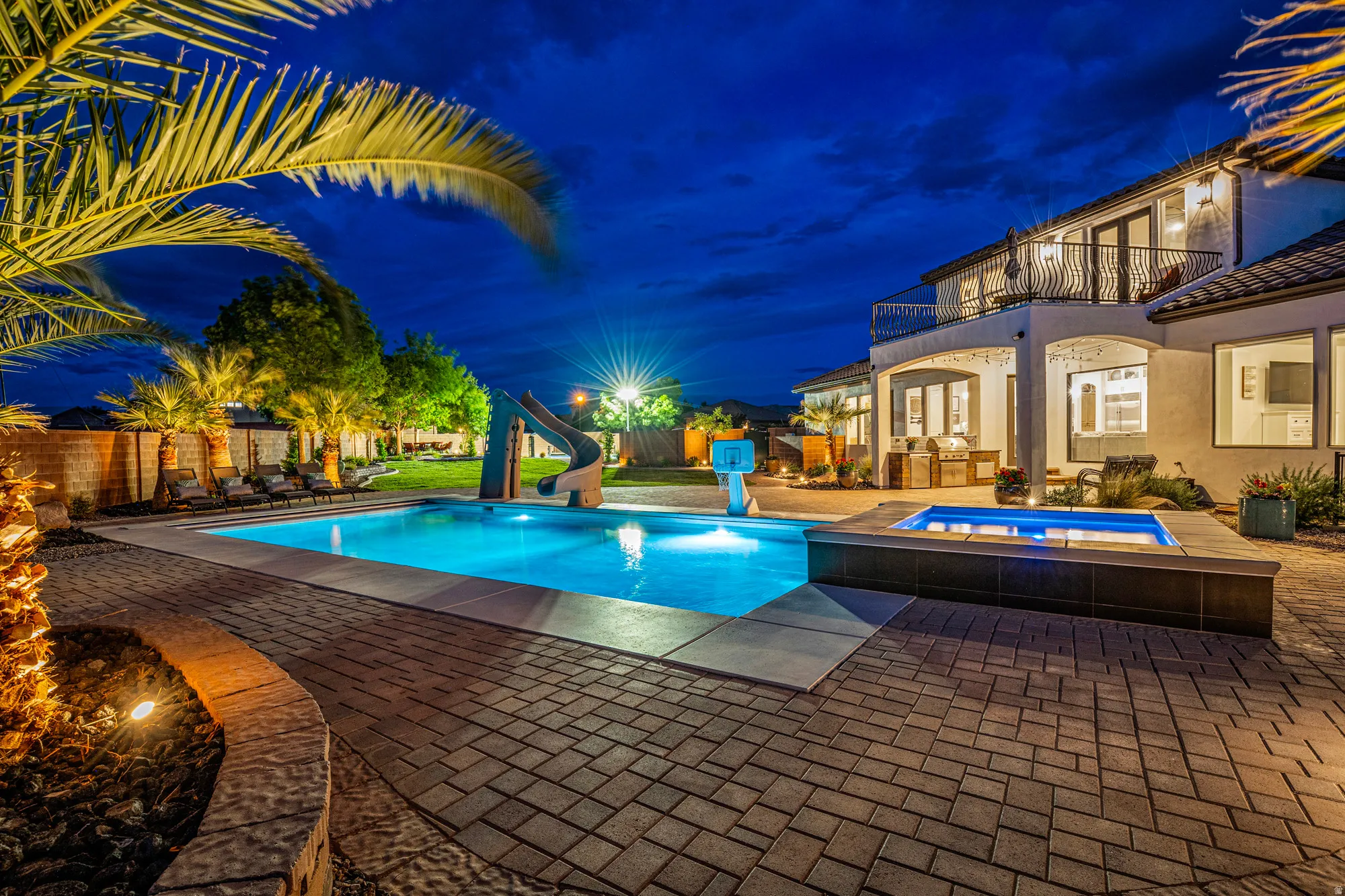 Expansive backyard featuring a large swimming pool with an integrated spa, paver patio, mature palm trees, and a second-story balcony