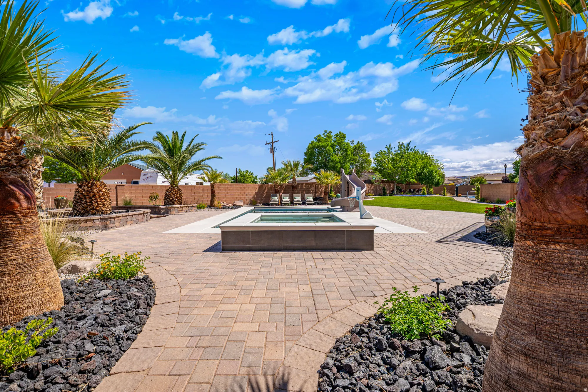 Expansive outdoor living area featuring a central pool/spa, paver patio, lush green lawn, mature palm trees, and decorative landscape rock