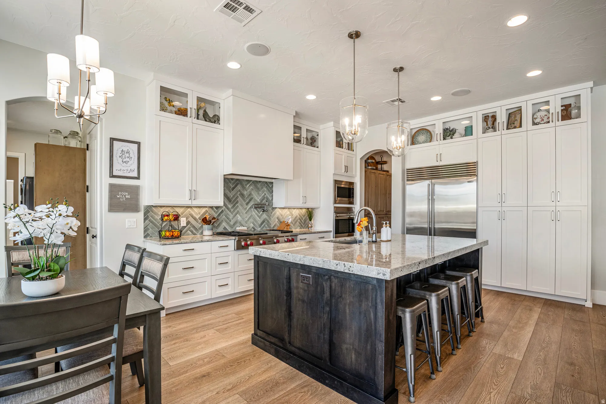 Spacious kitchen featuring an oversized island with seating, light wood-finish flooring, white cabinetry with glass upper doors, stainless steel appliances, and a herringbone tile backsplash